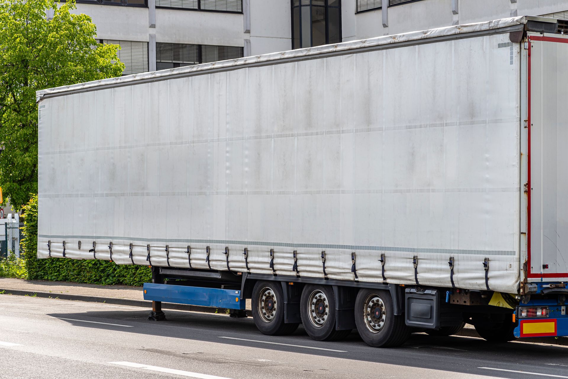 White semi-trailer parked on a street with triple axles, blue support frame, and a building in the background.