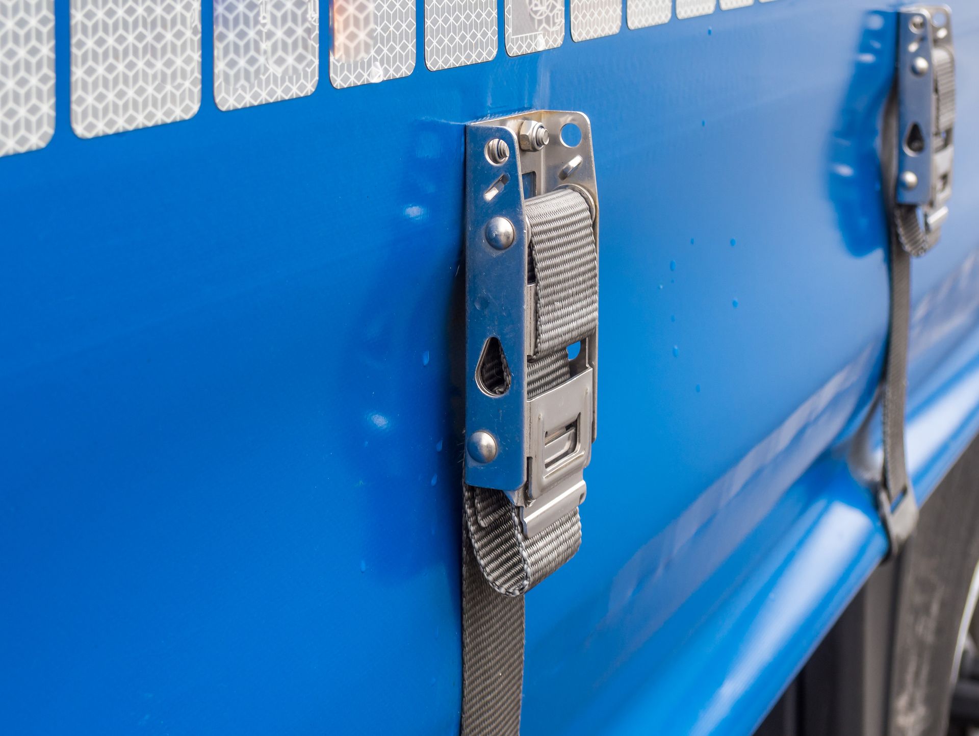 Close-up of a blue truck side with a metal buckle and grey strap for securing cargo.
