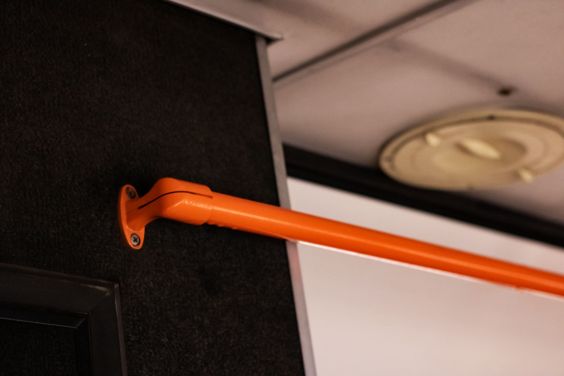 Orange handrail on a black wall inside a bus or train. Ceiling with light fixture in background.