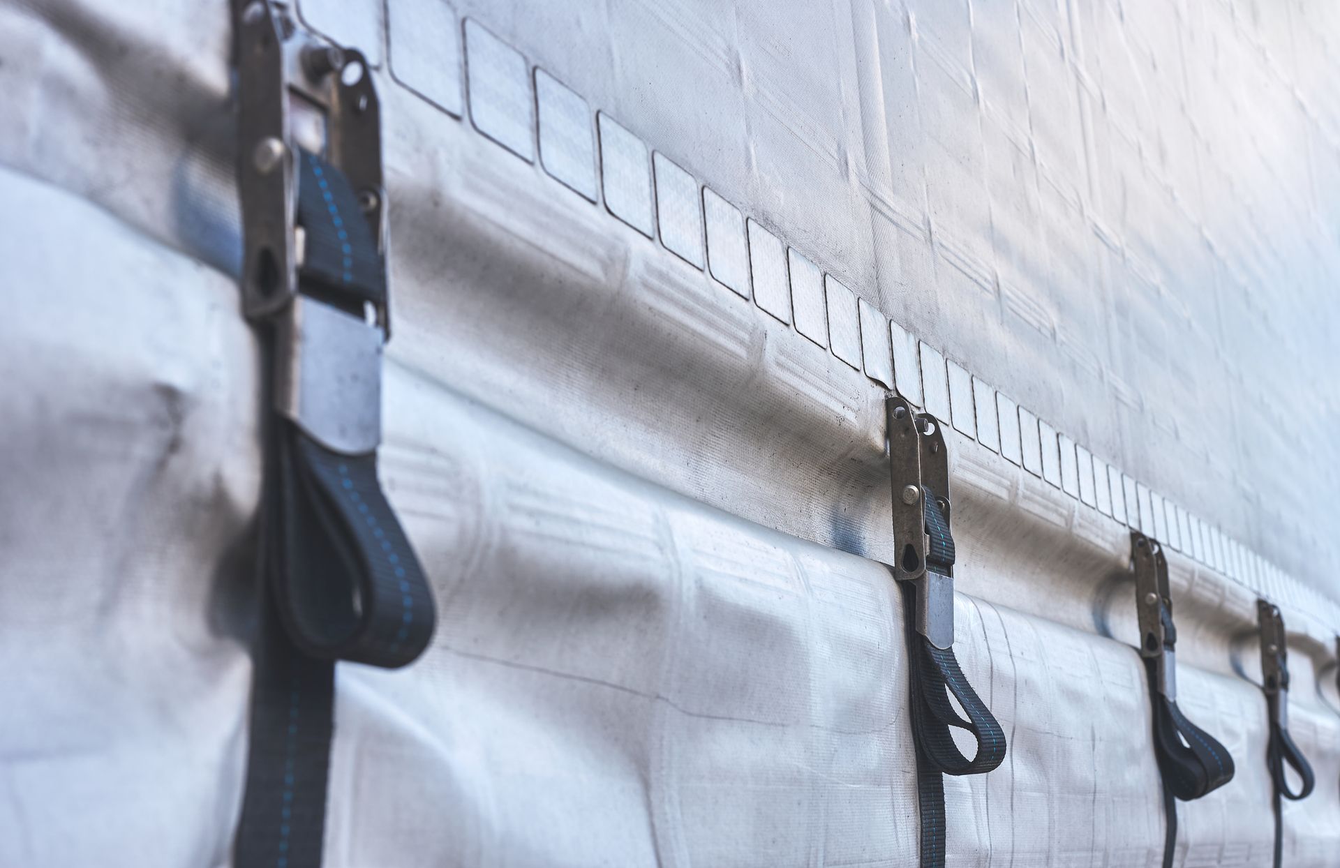 Close-up of a truck's side. Black straps secure the white tarp to the metallic frame with frequent square holes.