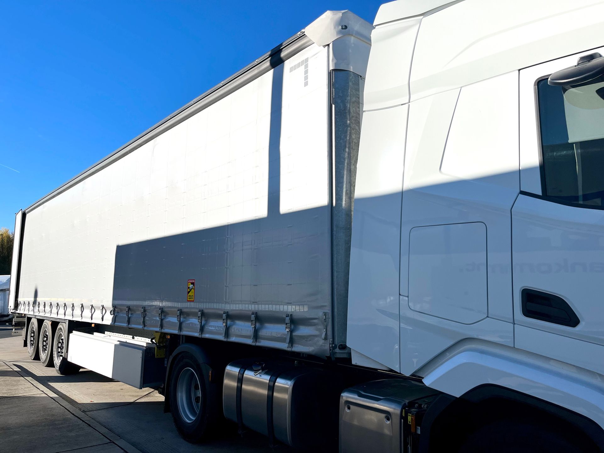 White semi-truck with a trailer on a sunny day. The trailer's side is white and has a metal undercarriage.