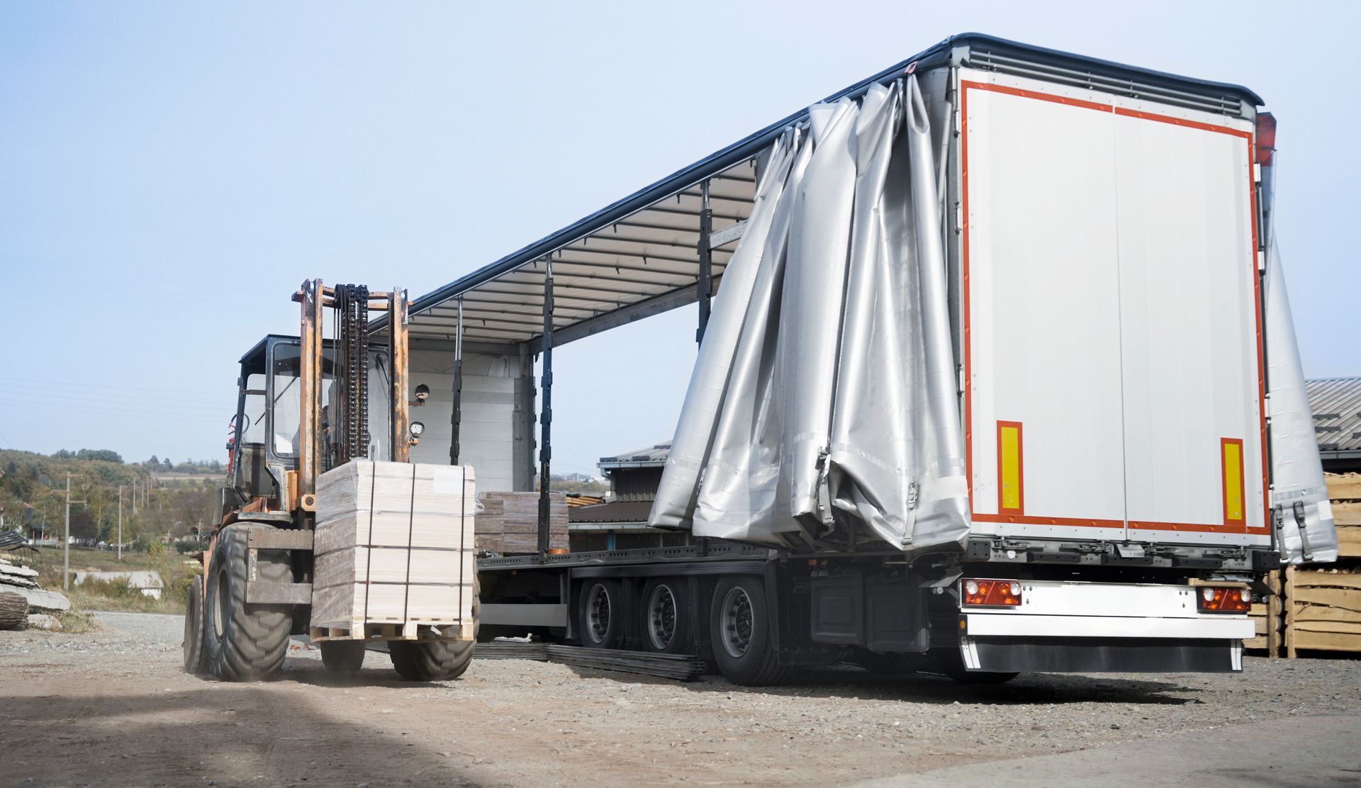 Forklift loading blocks onto an open-sided trailer in a gravel lot.