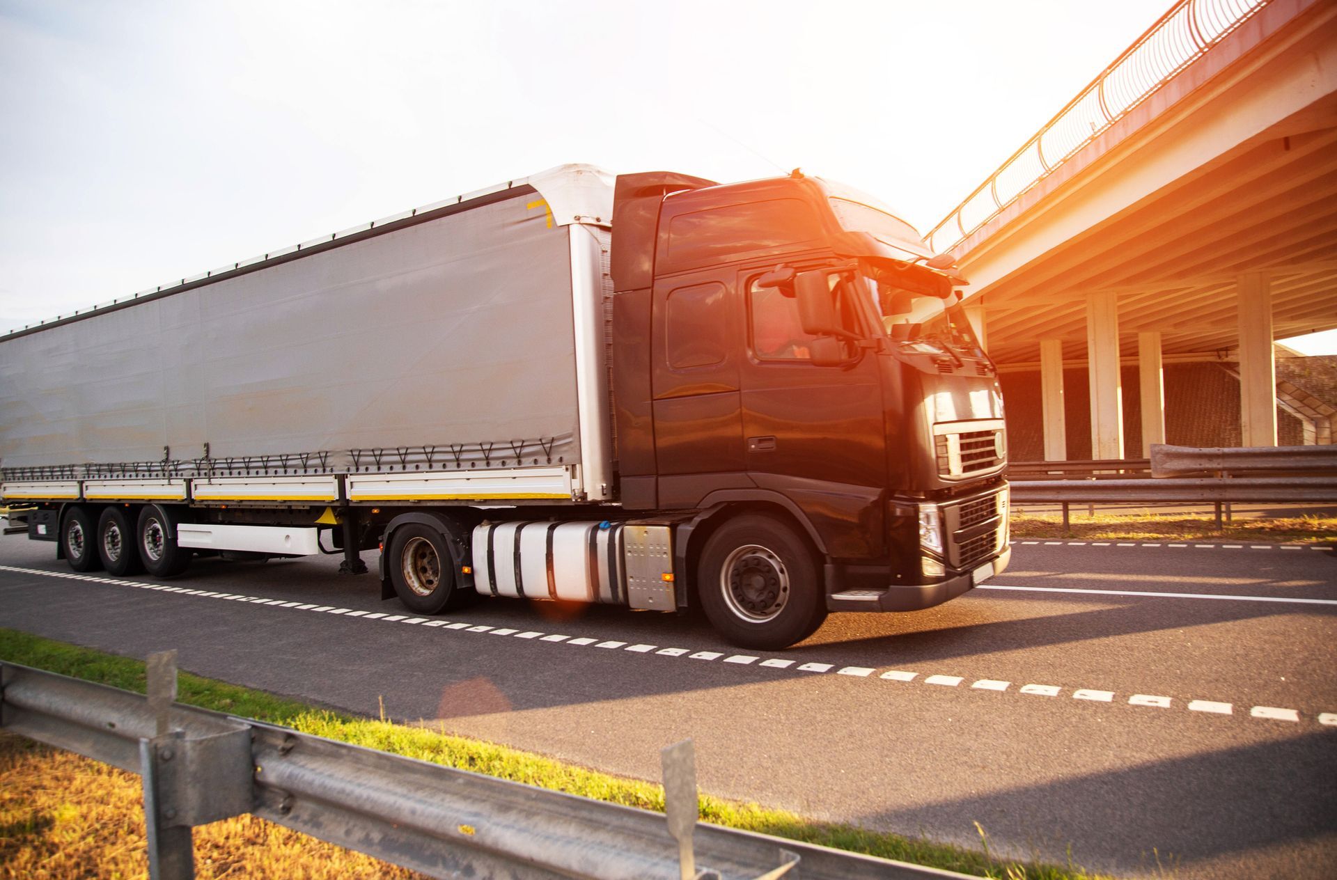 Black semi-truck driving on a highway near a bridge on a sunny day.