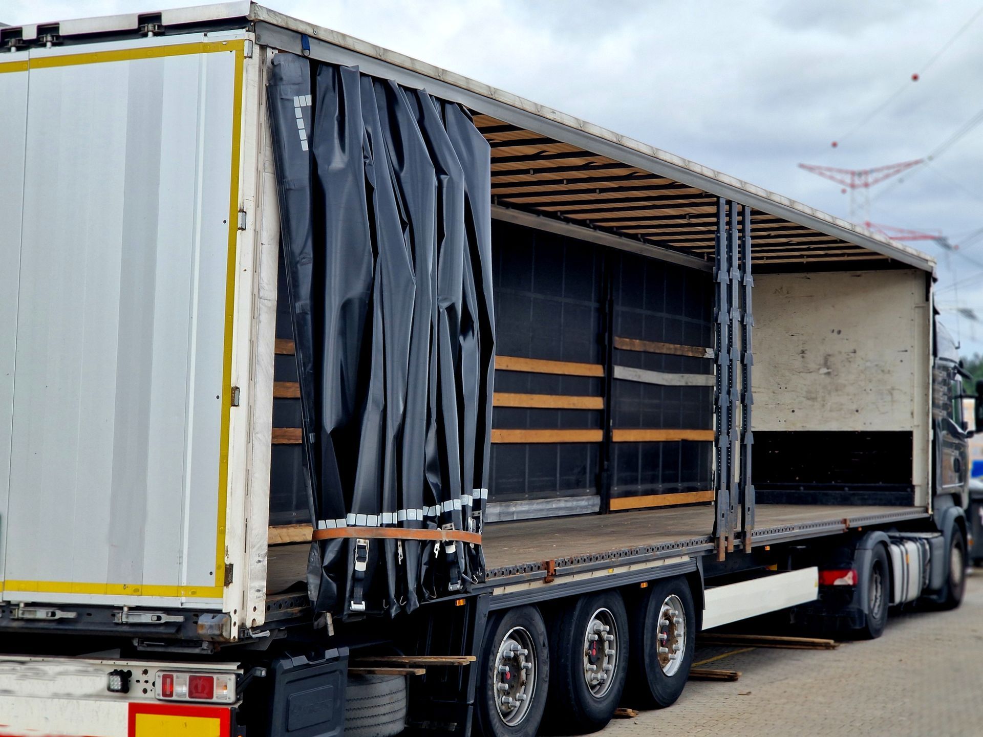 Open semi-trailer truck, black tarp partially covering the side, parked outdoors on a cloudy day.