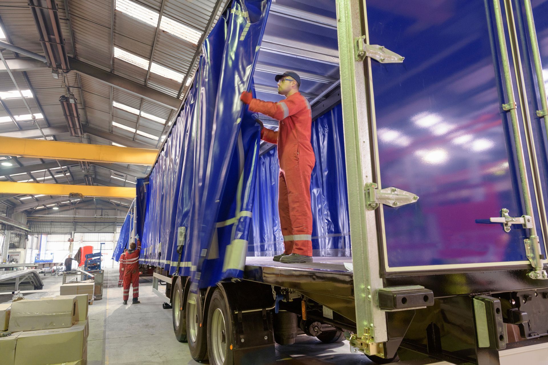 Workers in red coveralls securing a blue tarp on a semi-trailer inside a warehouse.