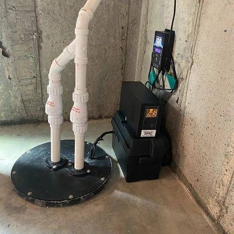 Basement sump pump system with white PVC pipes, black basin and battery backup, against concrete walls.