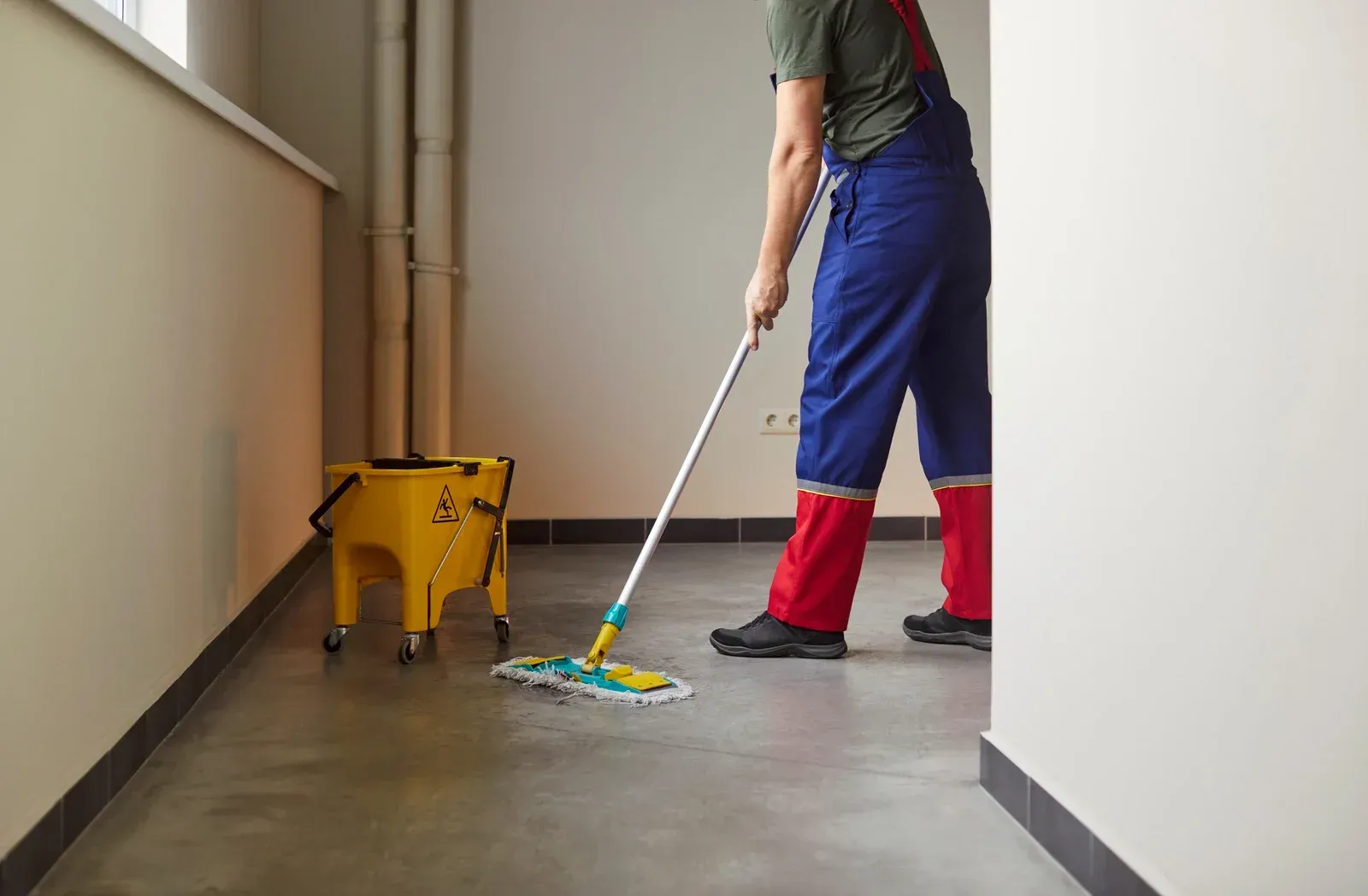 A person wearing blue and red work overalls mops a grey floor next to a yellow mop bucket in a hallway.