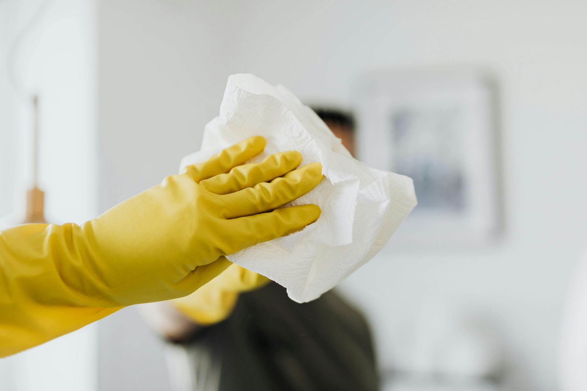 A hand in a yellow rubber glove holds a white cleaning cloth toward the camera in a blurred home interior.