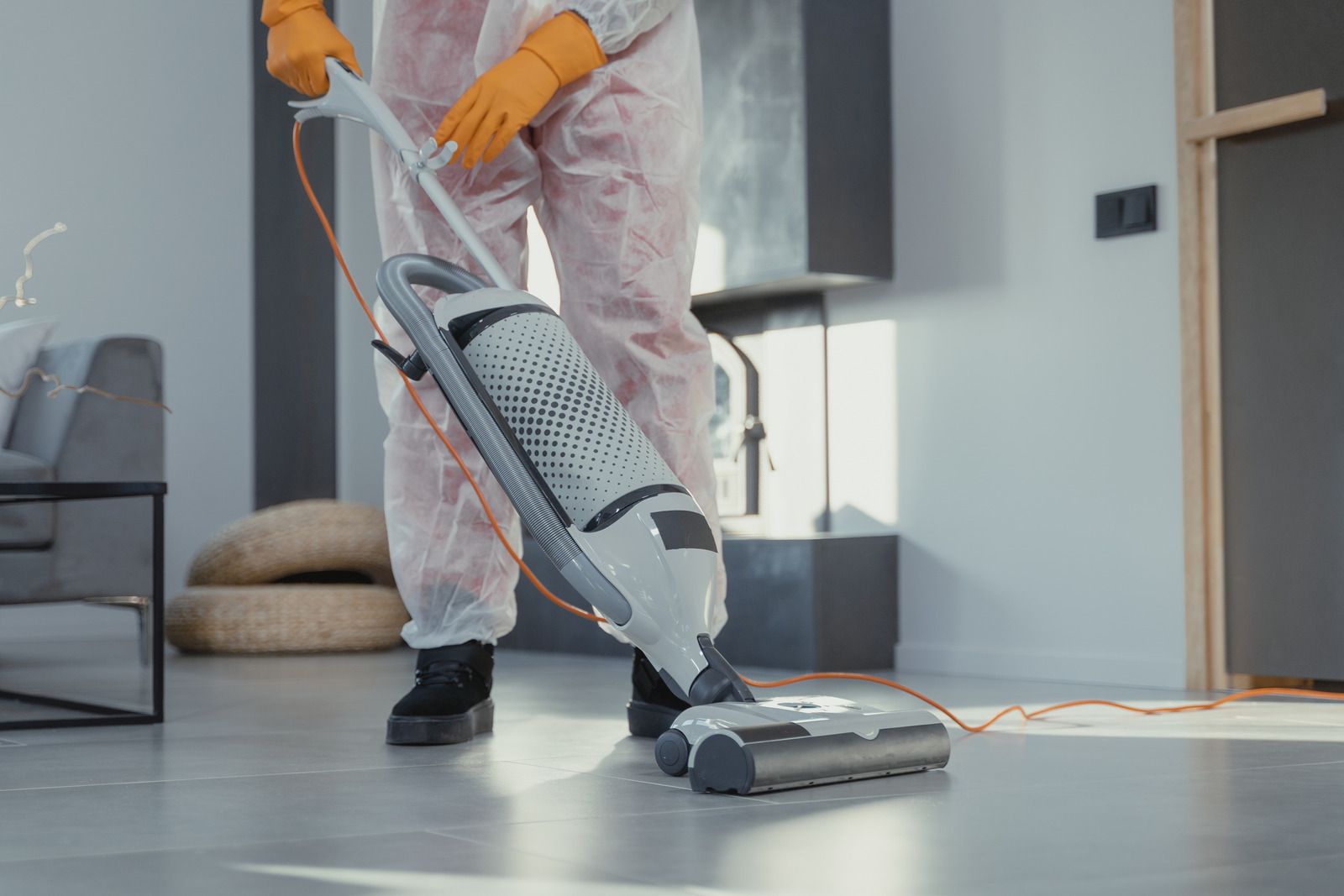 A person in protective white clothing and orange gloves vacuums a floor with an upright vacuum cleaner.