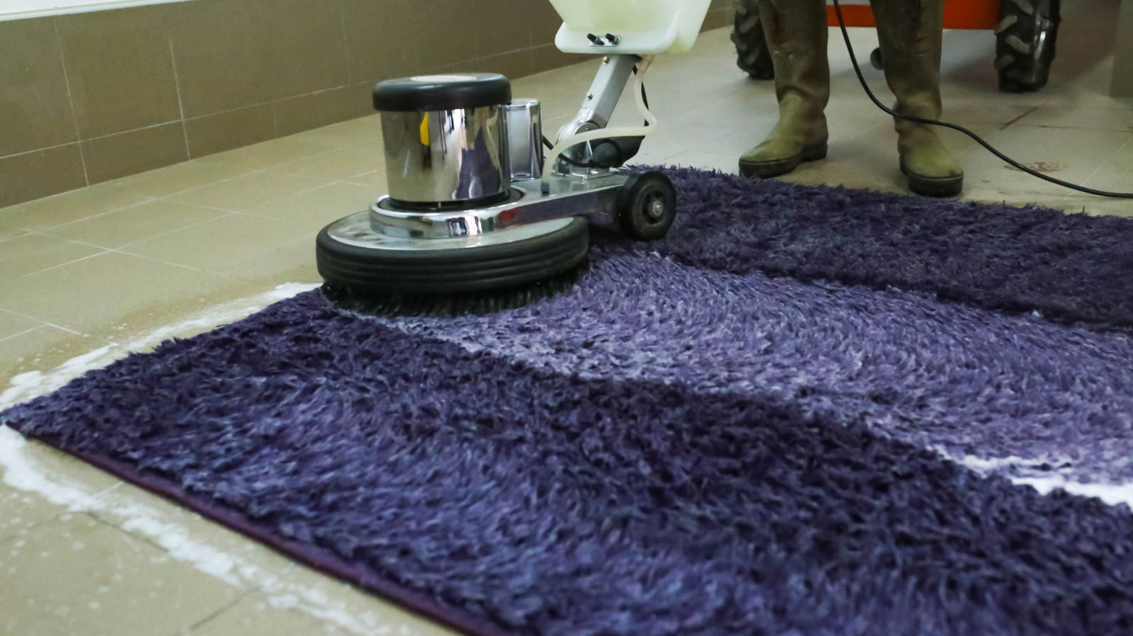 A person uses a floor-scrubbing machine to deep clean a shaggy purple rug in a tiled room.