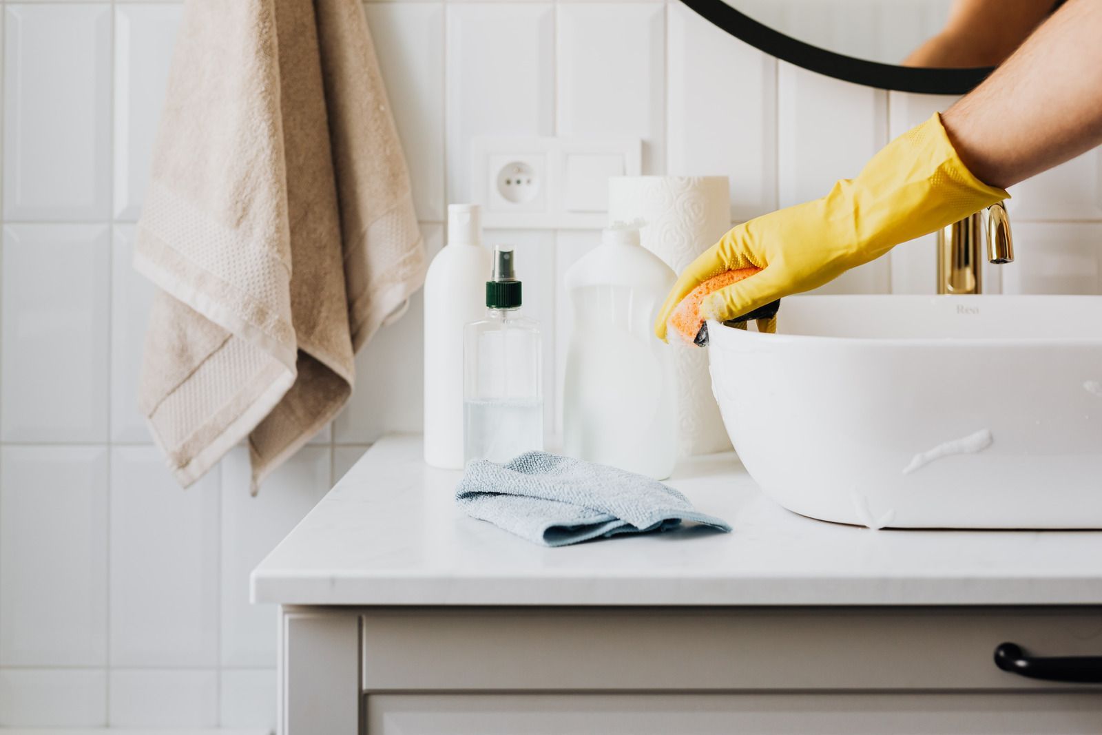 A hand wearing a yellow rubber glove cleans a white bathroom sink with a sponge, with cleaning supplies on the counter.