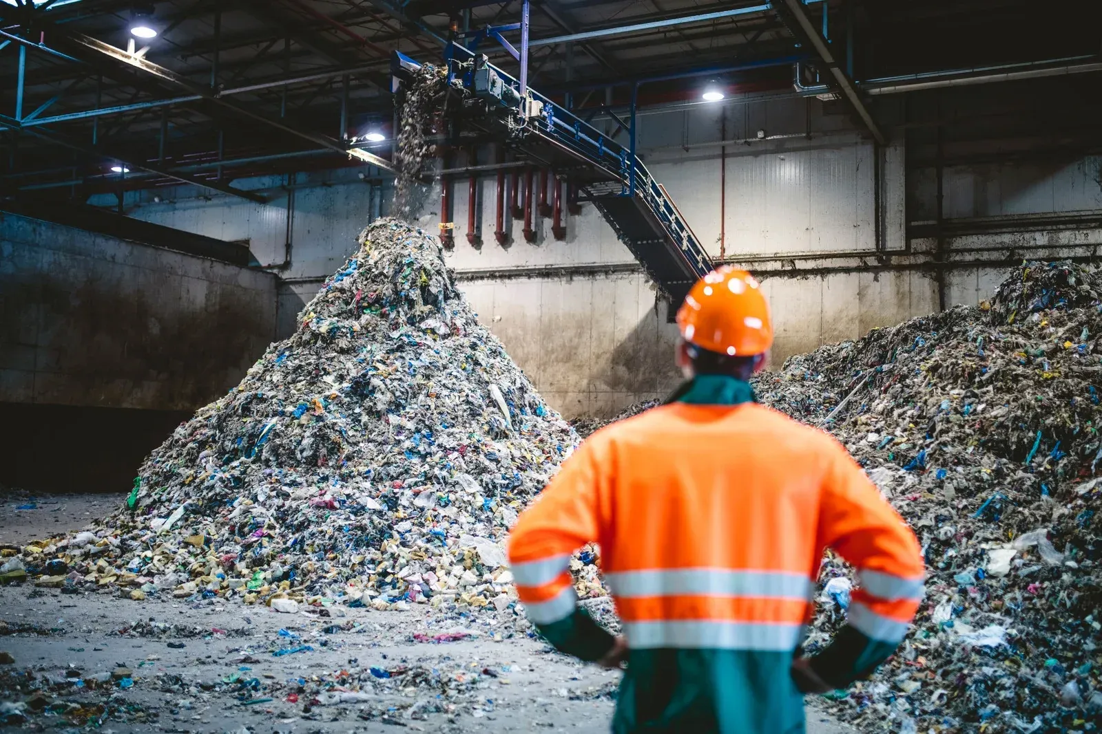 A worker in a high-visibility orange jacket and hard hat stands before large piles of waste in a recycling facility.