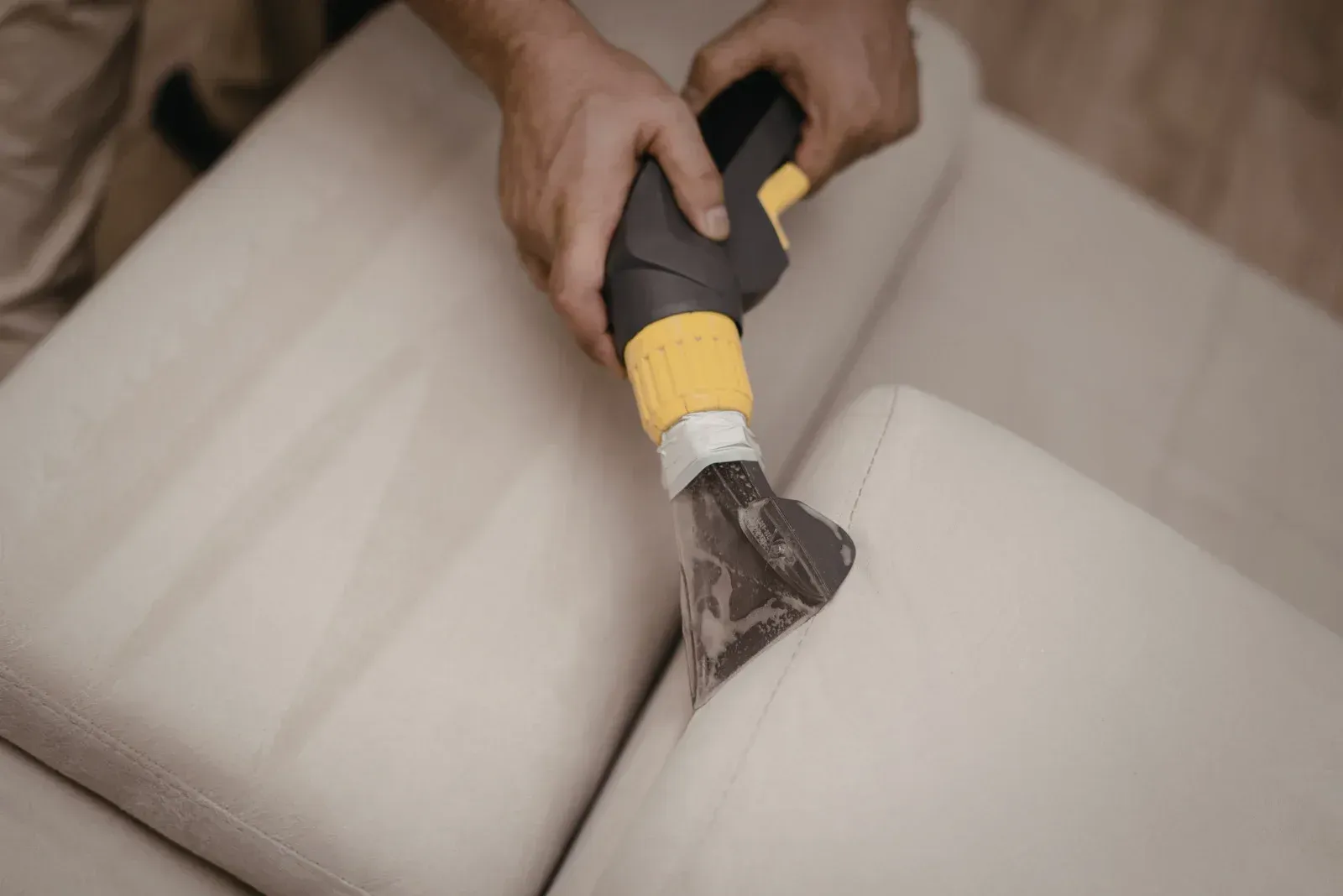 A person uses a handheld upholstery cleaner to deep clean a light-colored fabric sofa.