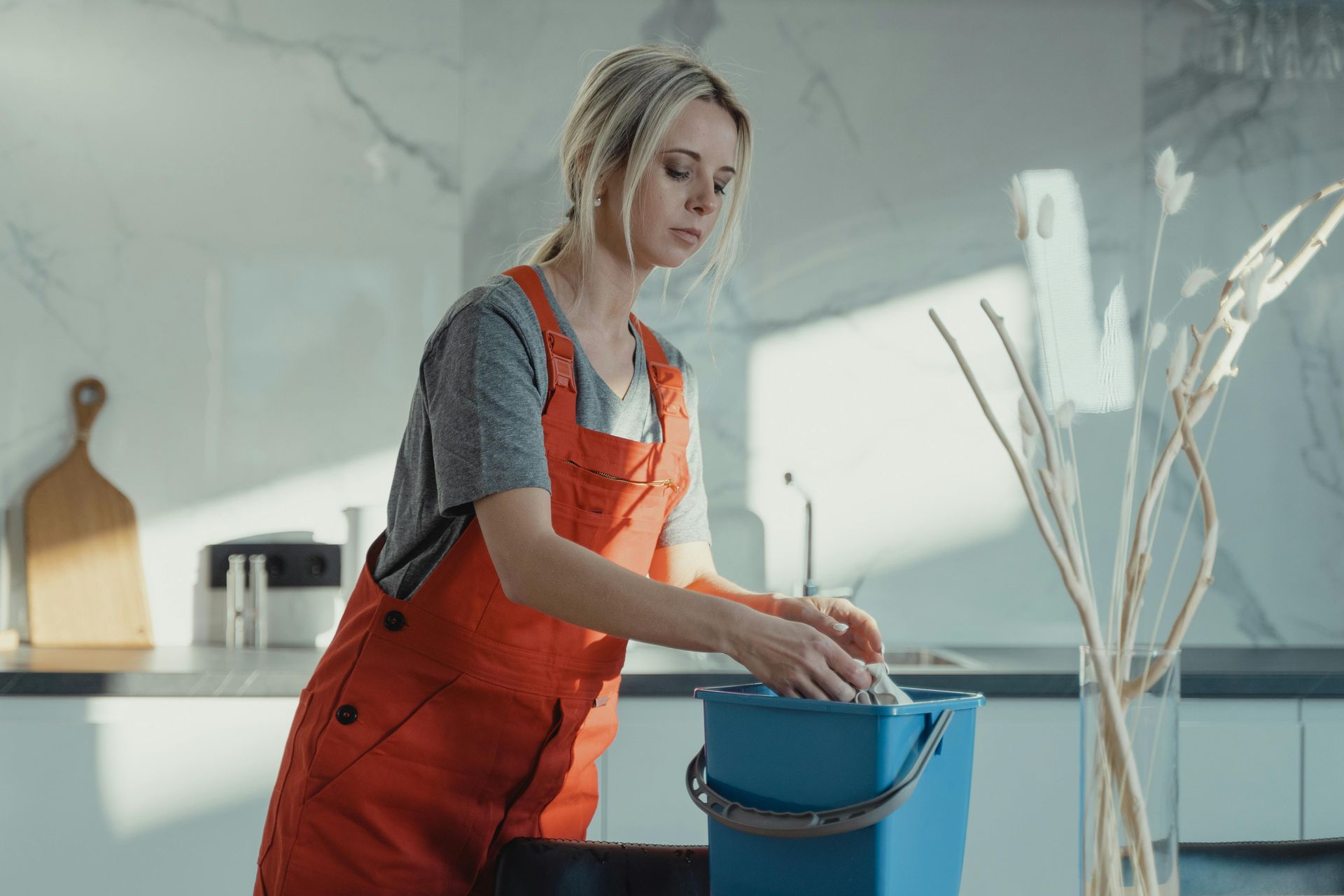 A person in bright orange overalls cleaning a mop in a blue bucket inside a sunlit kitchen.