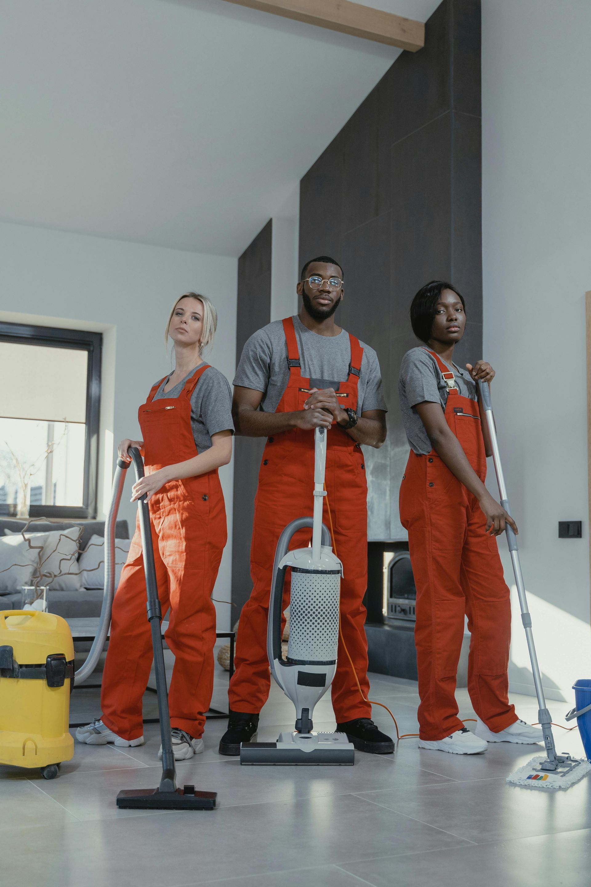 Three professionals in matching orange coveralls and grey shirts stand holding cleaning equipment in a modern room.