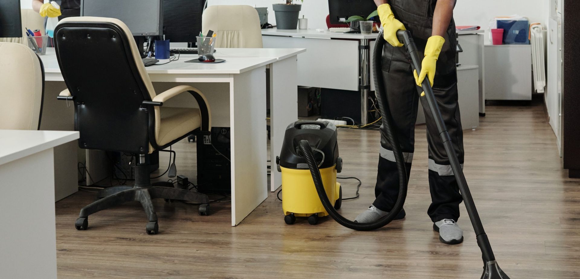 A person wearing yellow rubber gloves wipes down a black office chair on a wooden floor.
