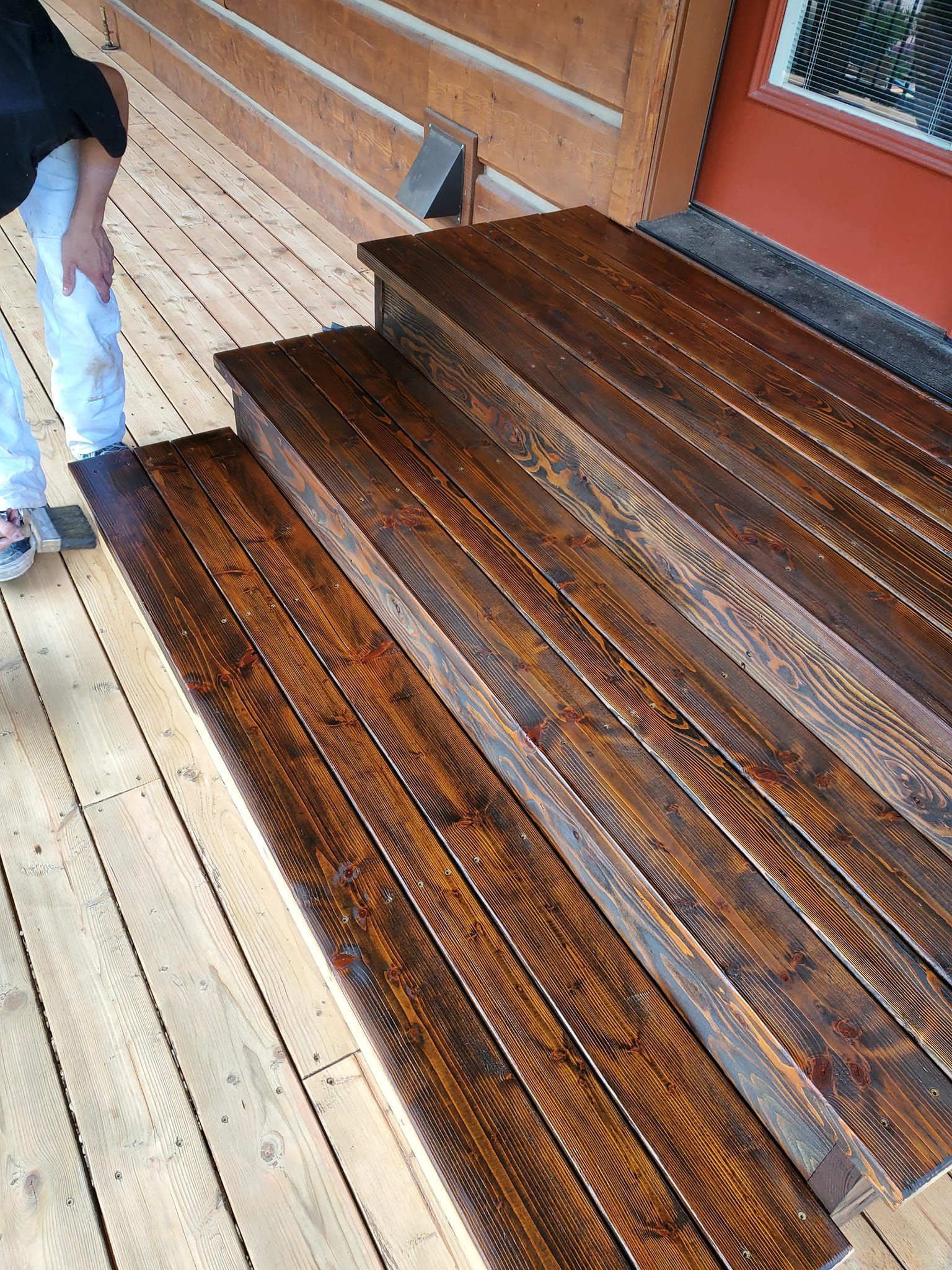 Wooden steps stained dark brown leading up to a door. A person stands nearby, touching their leg.