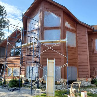House exterior being painted, scaffolding in place. Windows covered, brown siding, cloudy sky.