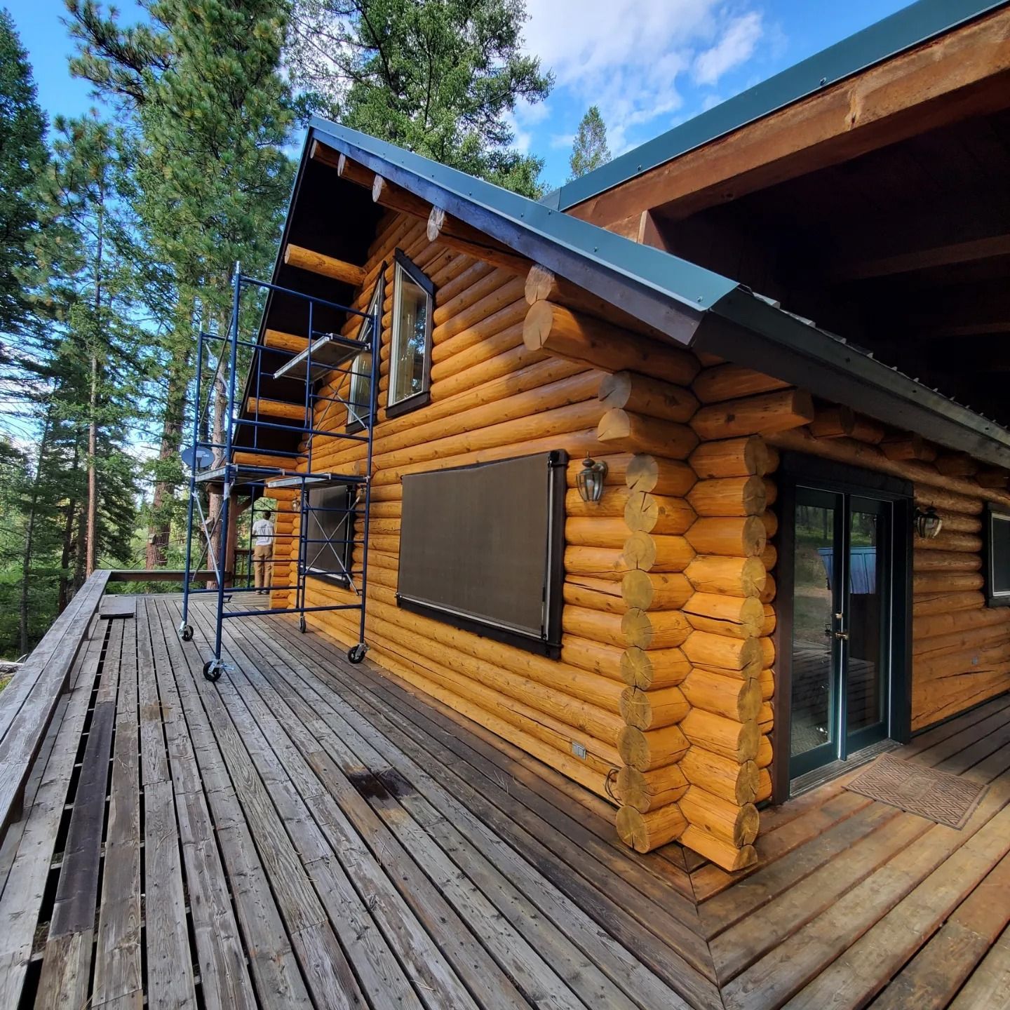 Log cabin with wooden deck, surrounded by trees. Scaffolding is set up on the side.