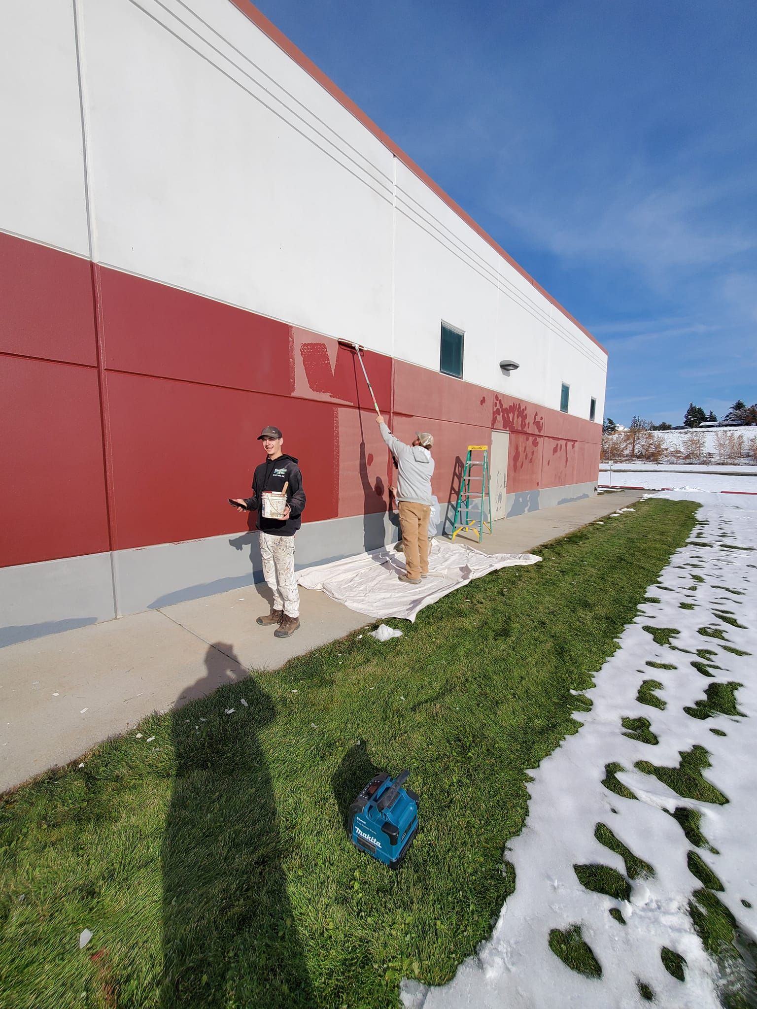 Two people painting the red exterior of a building. One uses a roller, the other holds a clipboard. Snow and grass nearby.