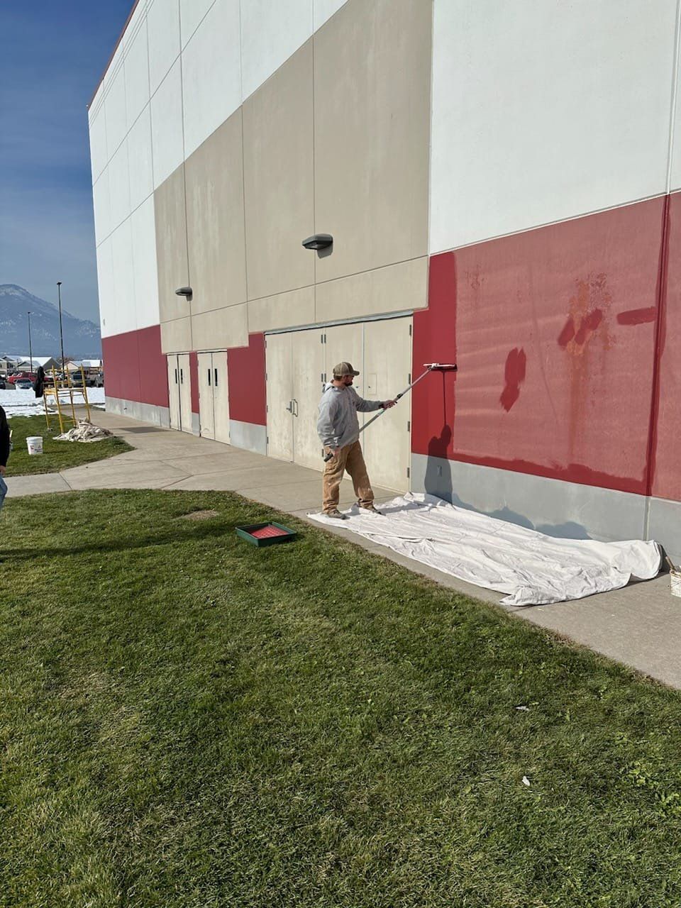 Person painting red section of building exterior with a roller. Building has white, tan, and red sections; outdoor setting.