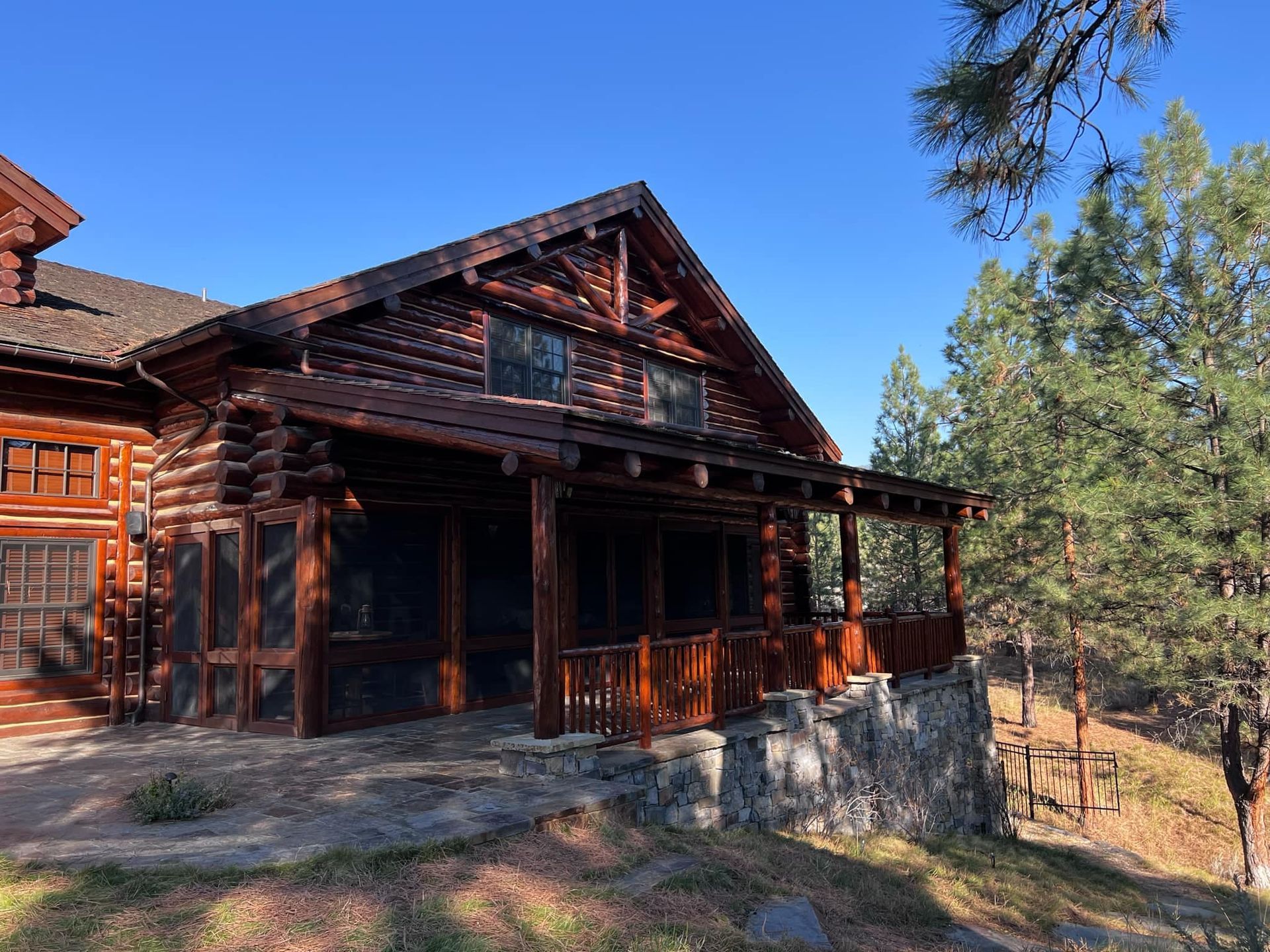 Log cabin with screened porch and timber roof under a clear blue sky, set in a wooded area.