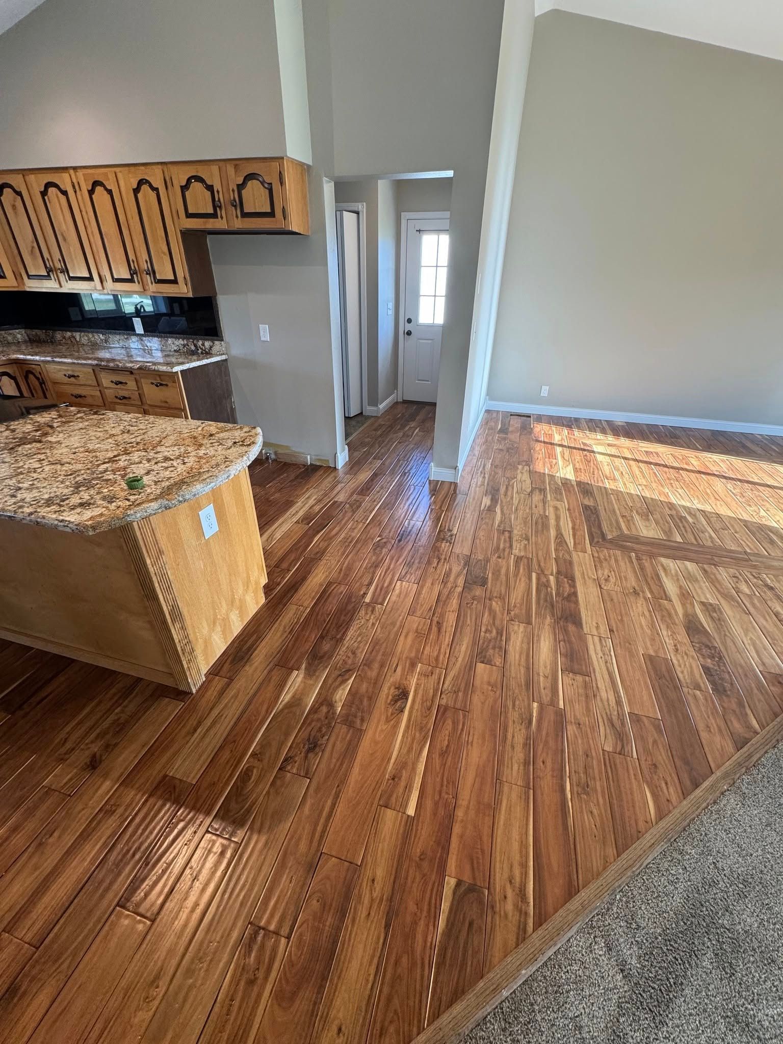 Hardwood floors in a kitchen, island with granite countertop. Wooden cabinets and a door are visible.