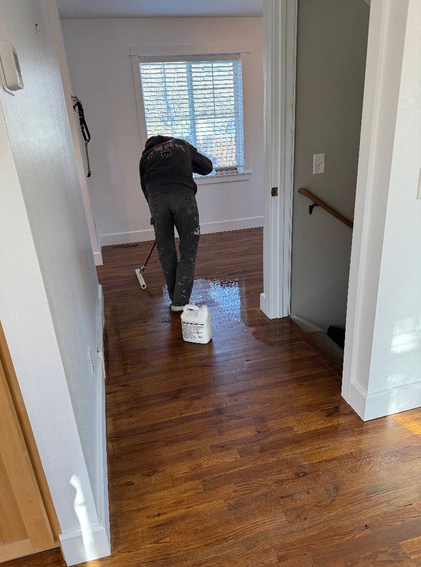 Person applying sealant to hardwood floor with roller, in a room with a window. Bottle of sealant on floor.