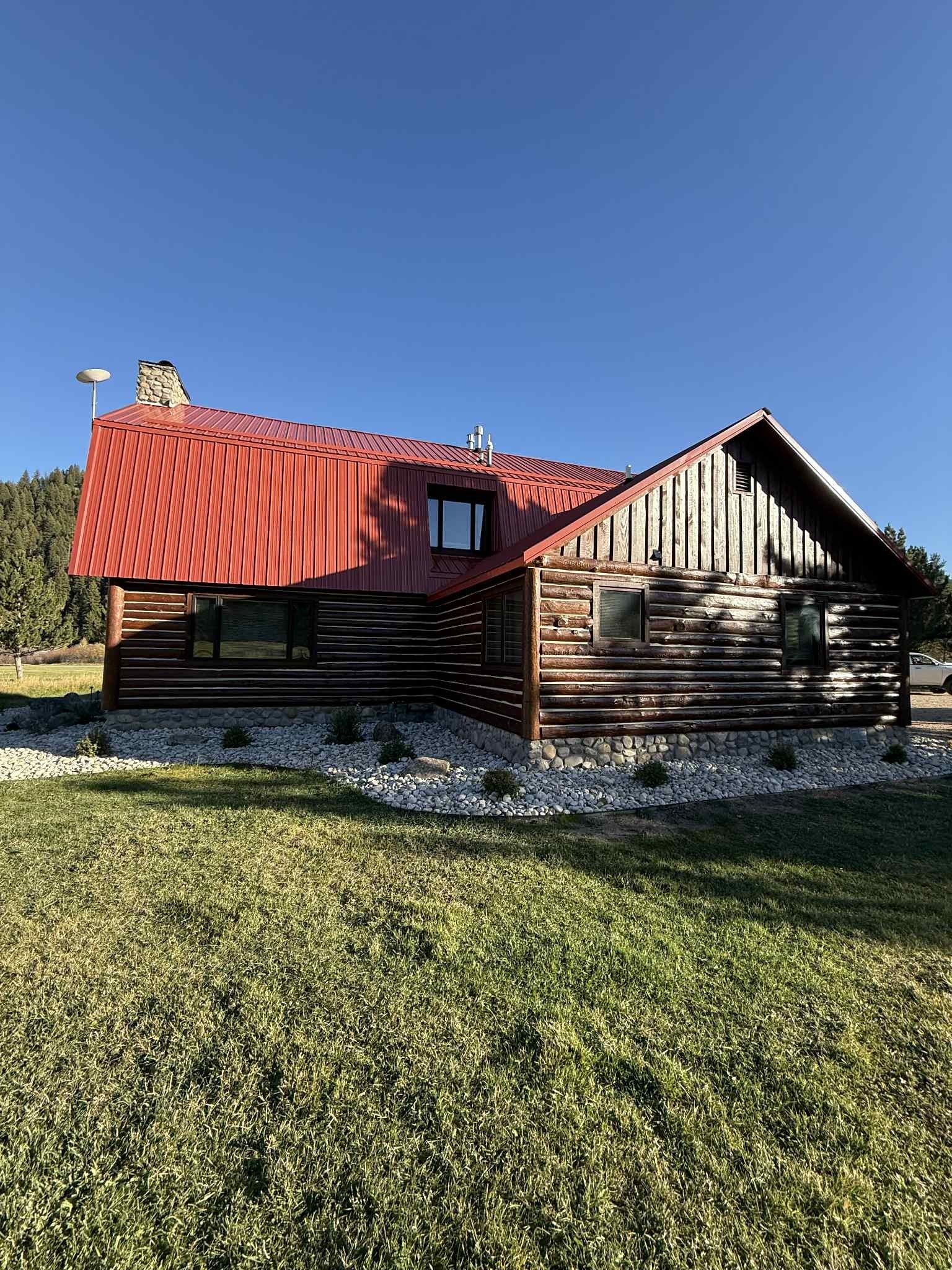 Log cabin with red metal roof under a blue sky, surrounded by green grass and rocks.