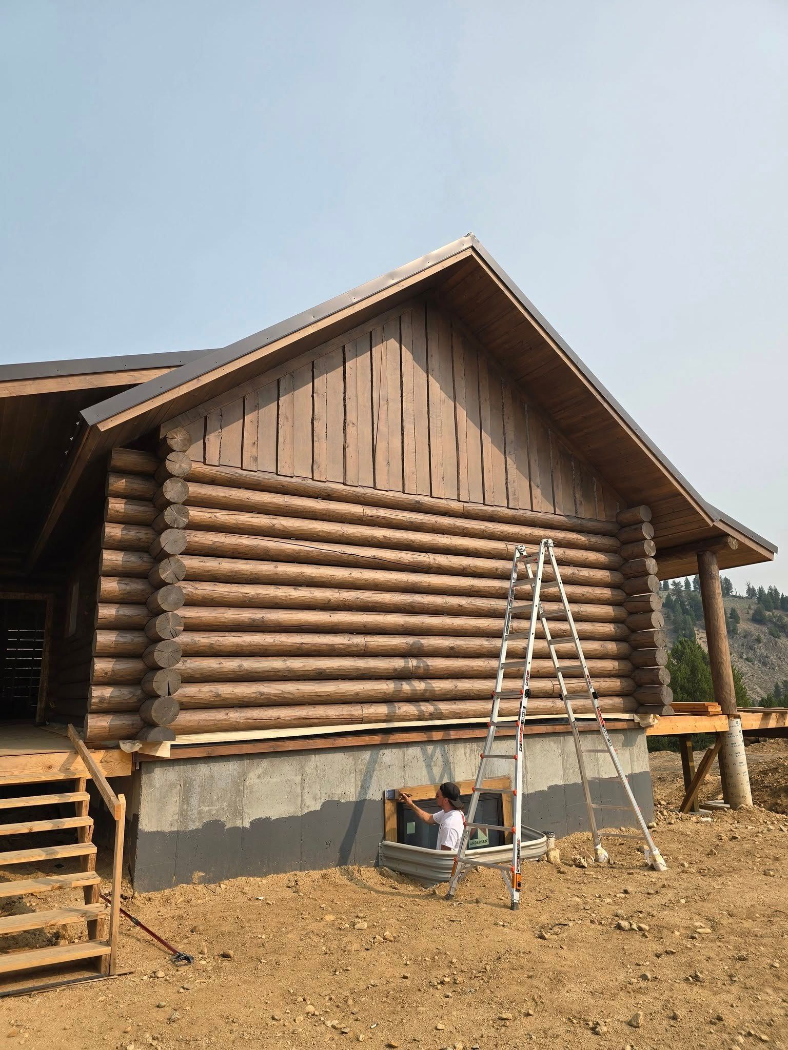 Wooden cabin under construction with a person working near a window; ladder present.