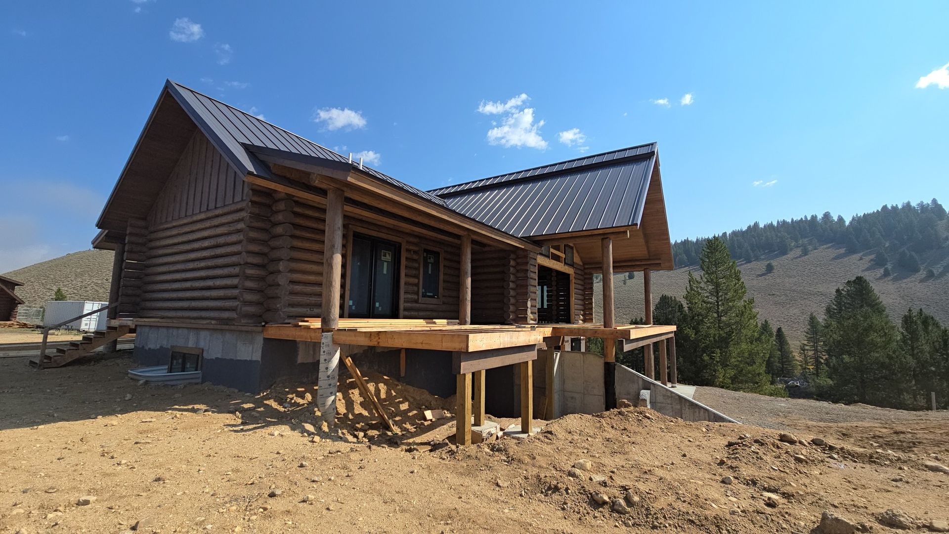Log cabin under construction with a deck, dark metal roof, and blue sky.