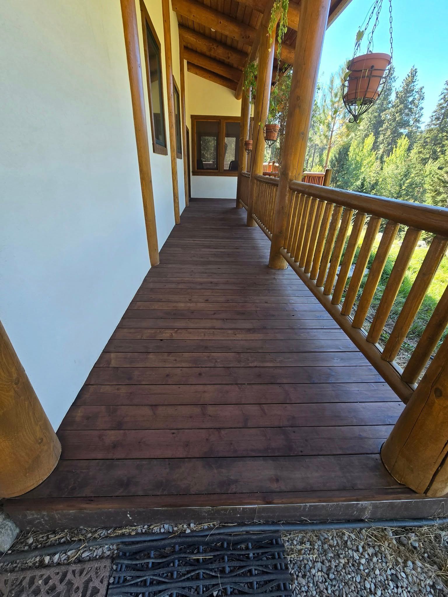Wooden porch with brown floorboards and railing, surrounded by wooden columns and a white wall, leading to a doorway.