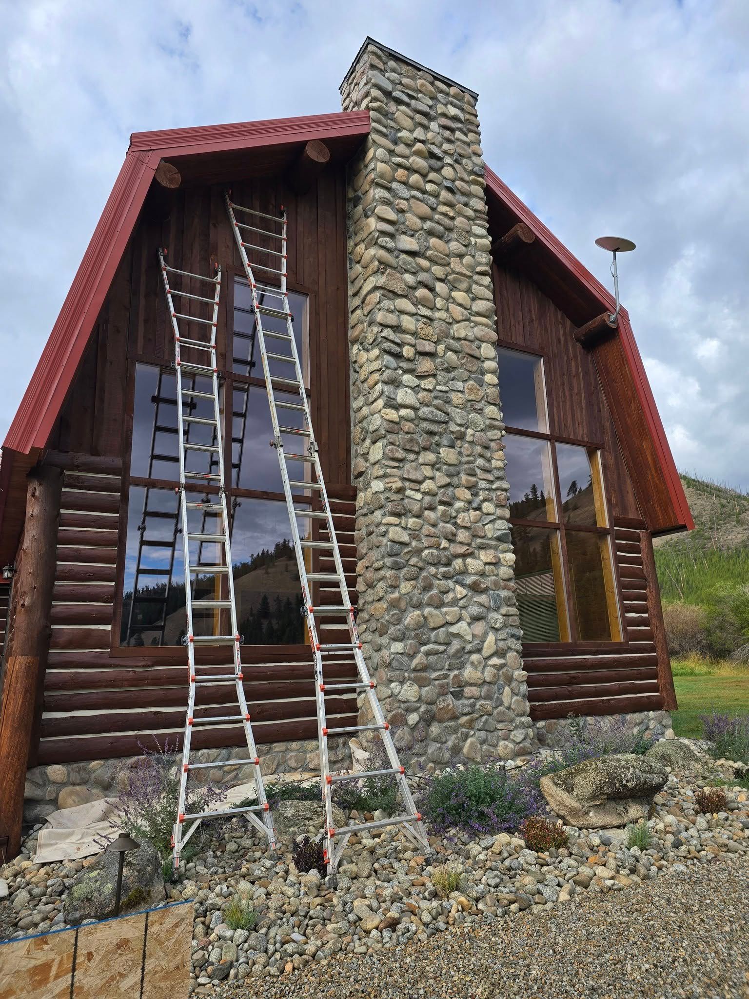 Two ladders lean against a cabin with a stone chimney. Cloudy sky. Brown logs and red roof.