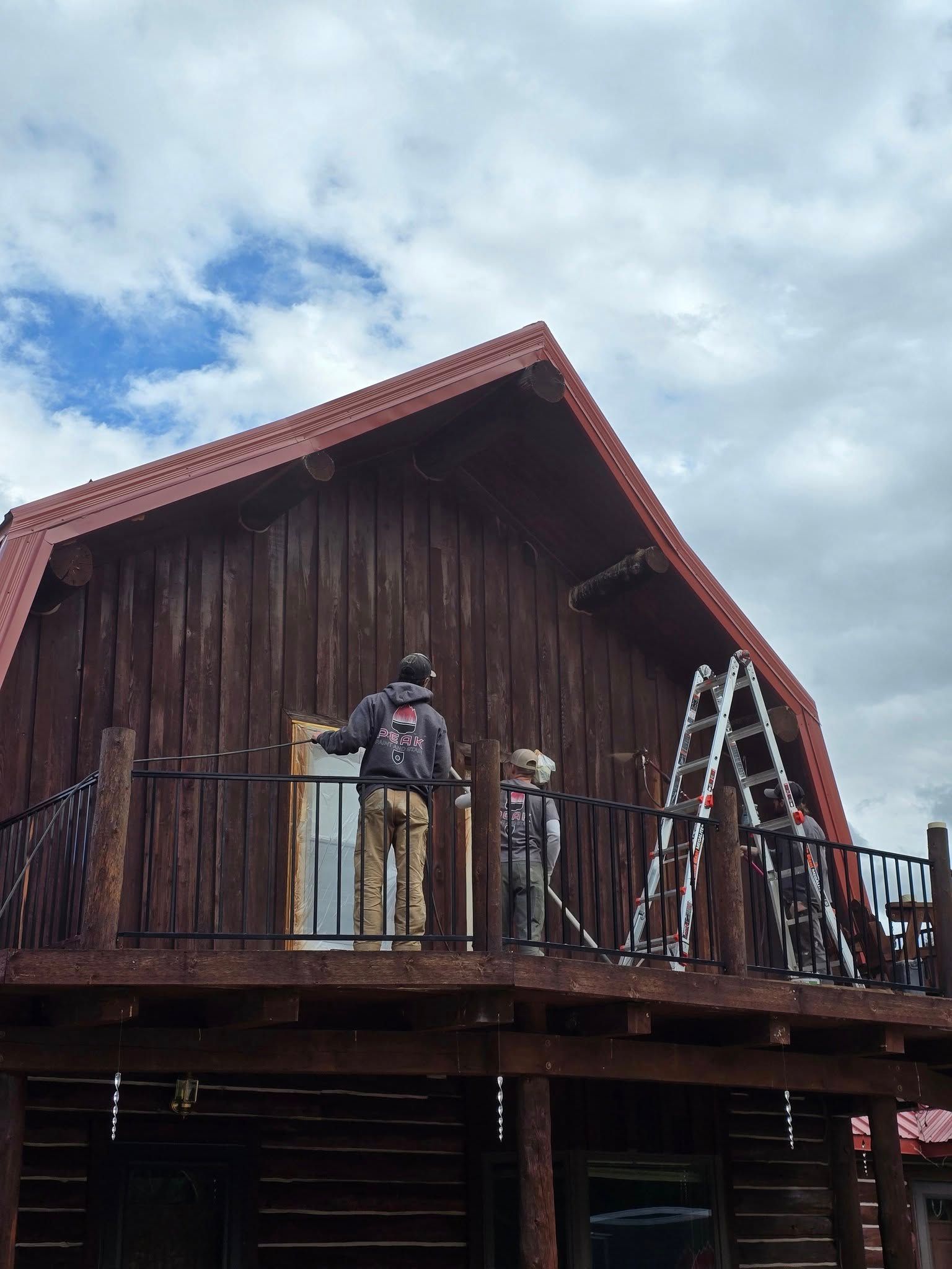 Two people on balcony of brown barn-style building, working with ladder and tools under cloudy sky.
