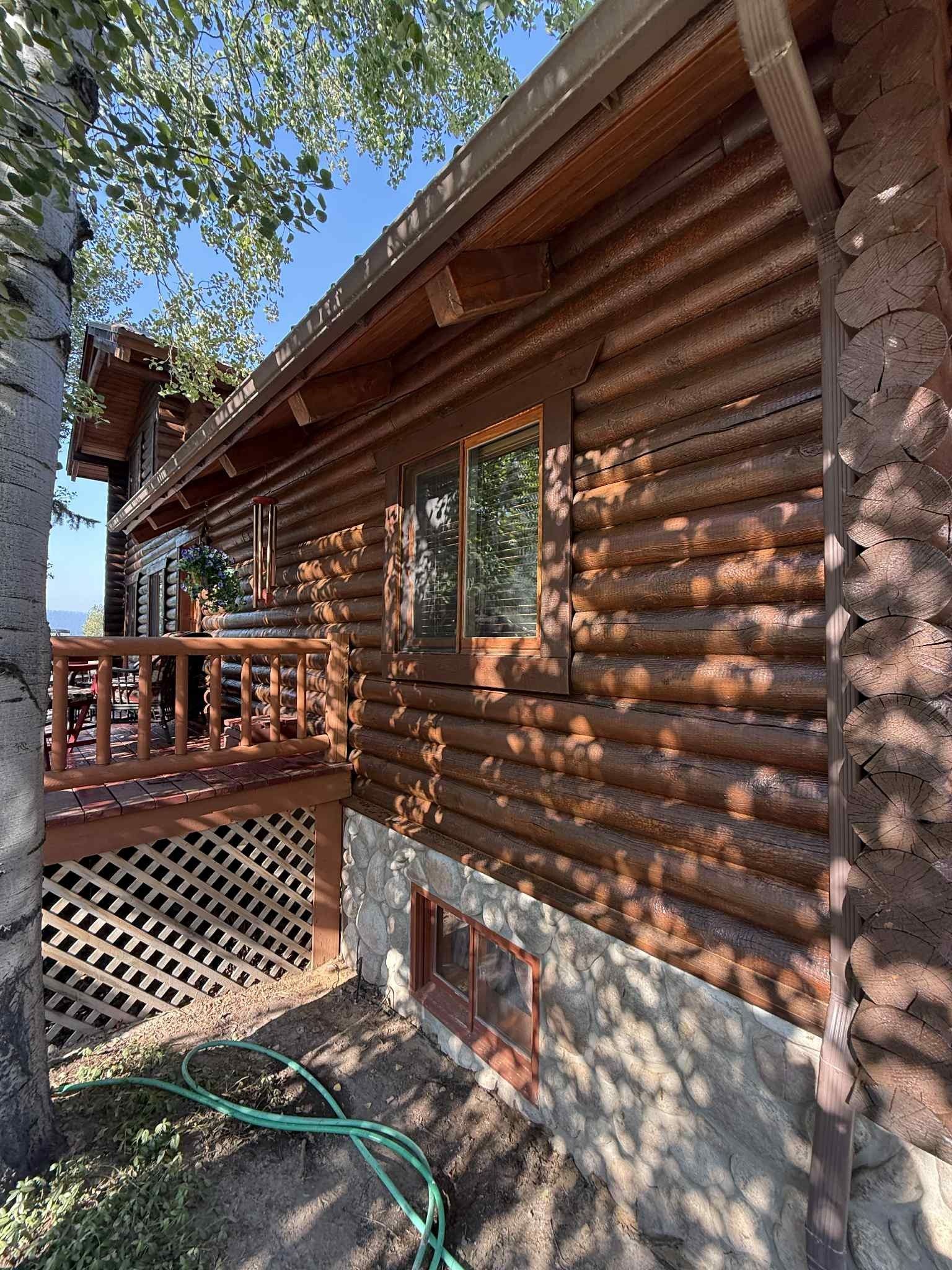 Log cabin with brown logs, window, and wooden deck. Green hose on ground, sunny day.