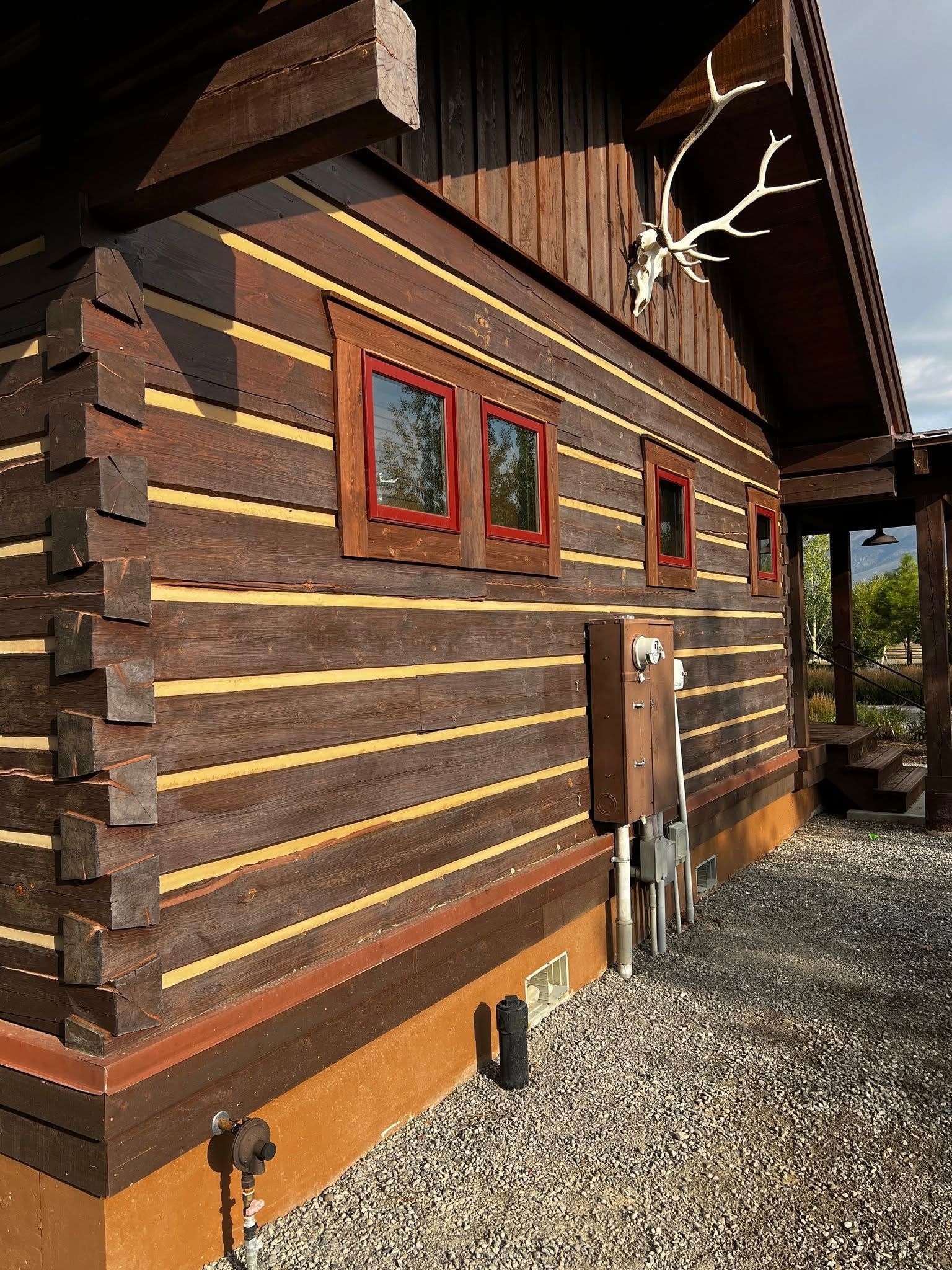 Log cabin exterior with brown stained logs, red window frames, and deer antlers mounted.
