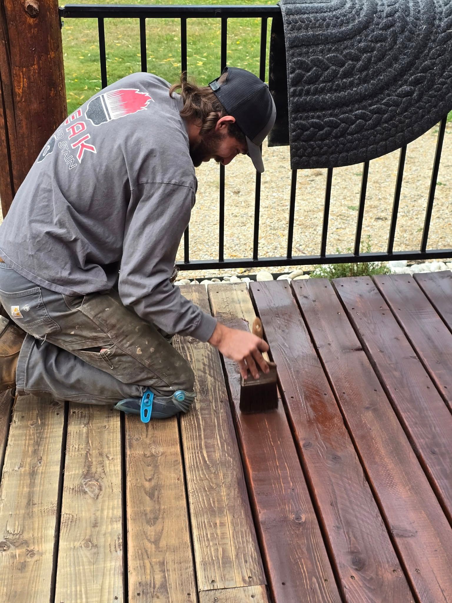 Person staining a wooden deck with a brush. Deck shows stained and unstained sections.