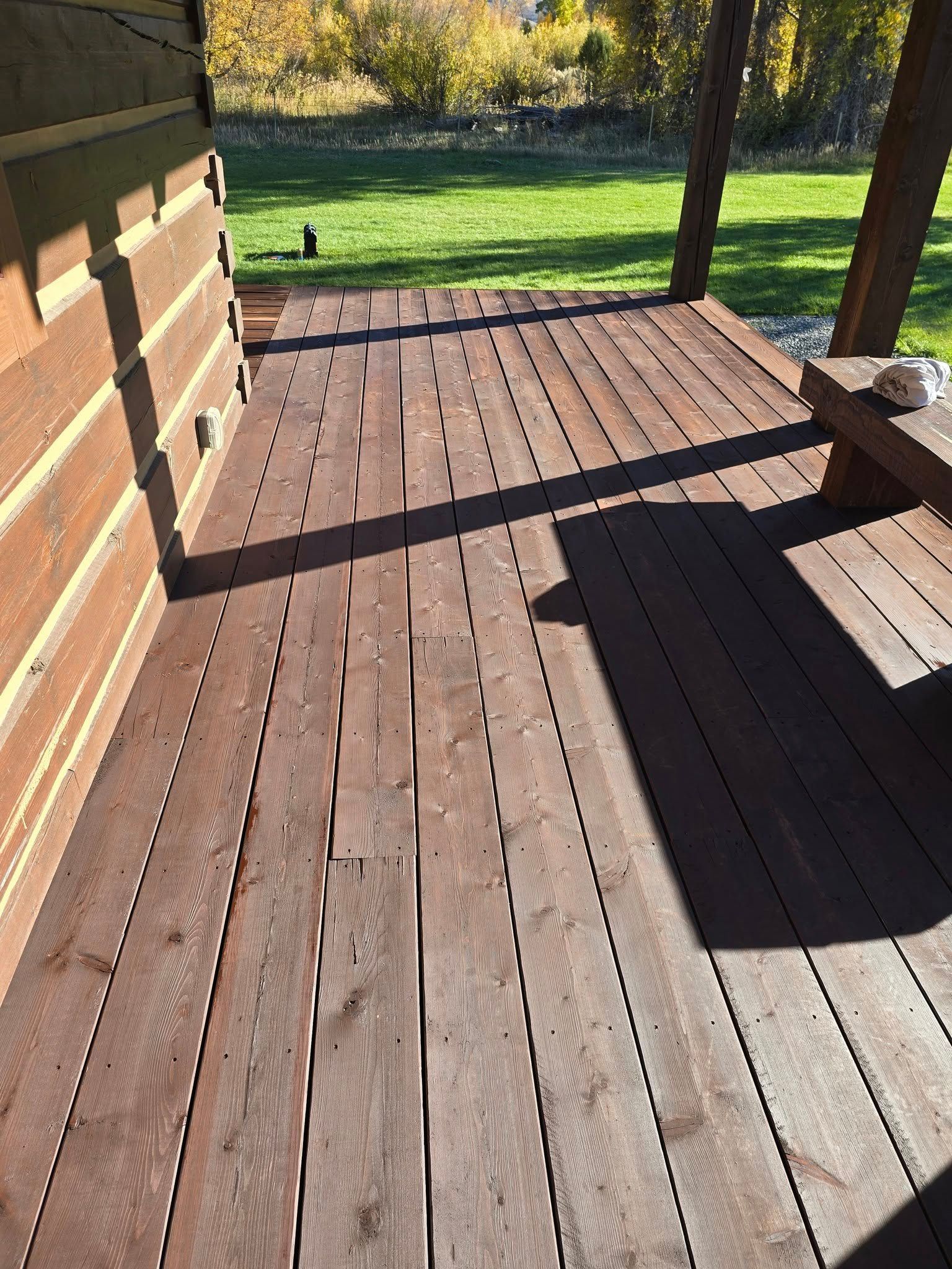 Wooden deck with reddish-brown planks, next to a log cabin, in a sunny outdoor setting.