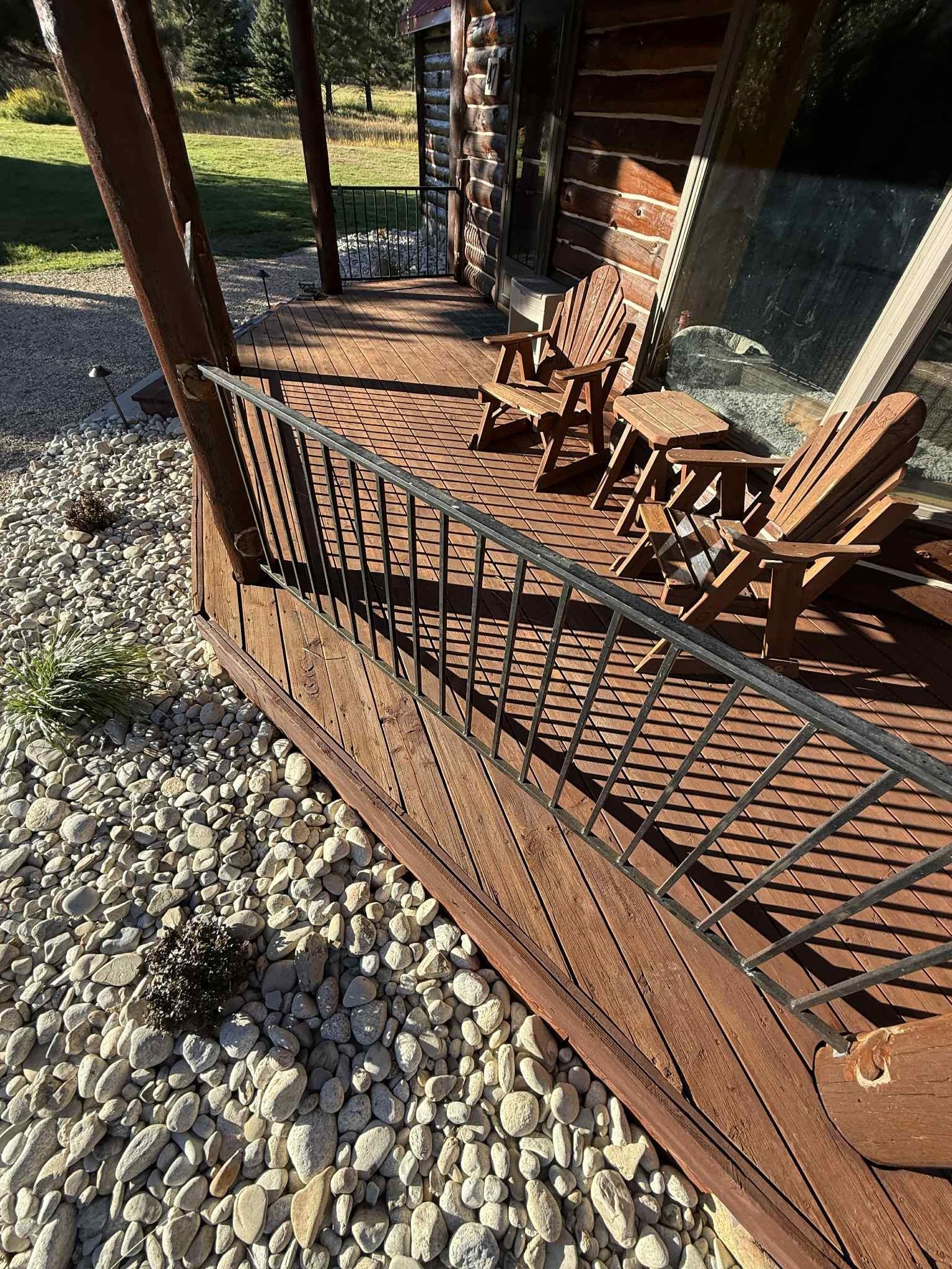 Wooden deck with railing, two chairs, and small table on a porch of a log cabin, surrounded by rocks.