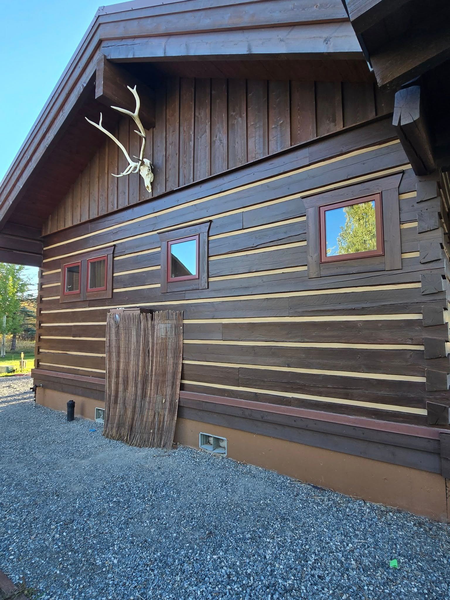 Wooden cabin exterior with antler mount and small windows. Dark brown logs and trim, gravel ground.