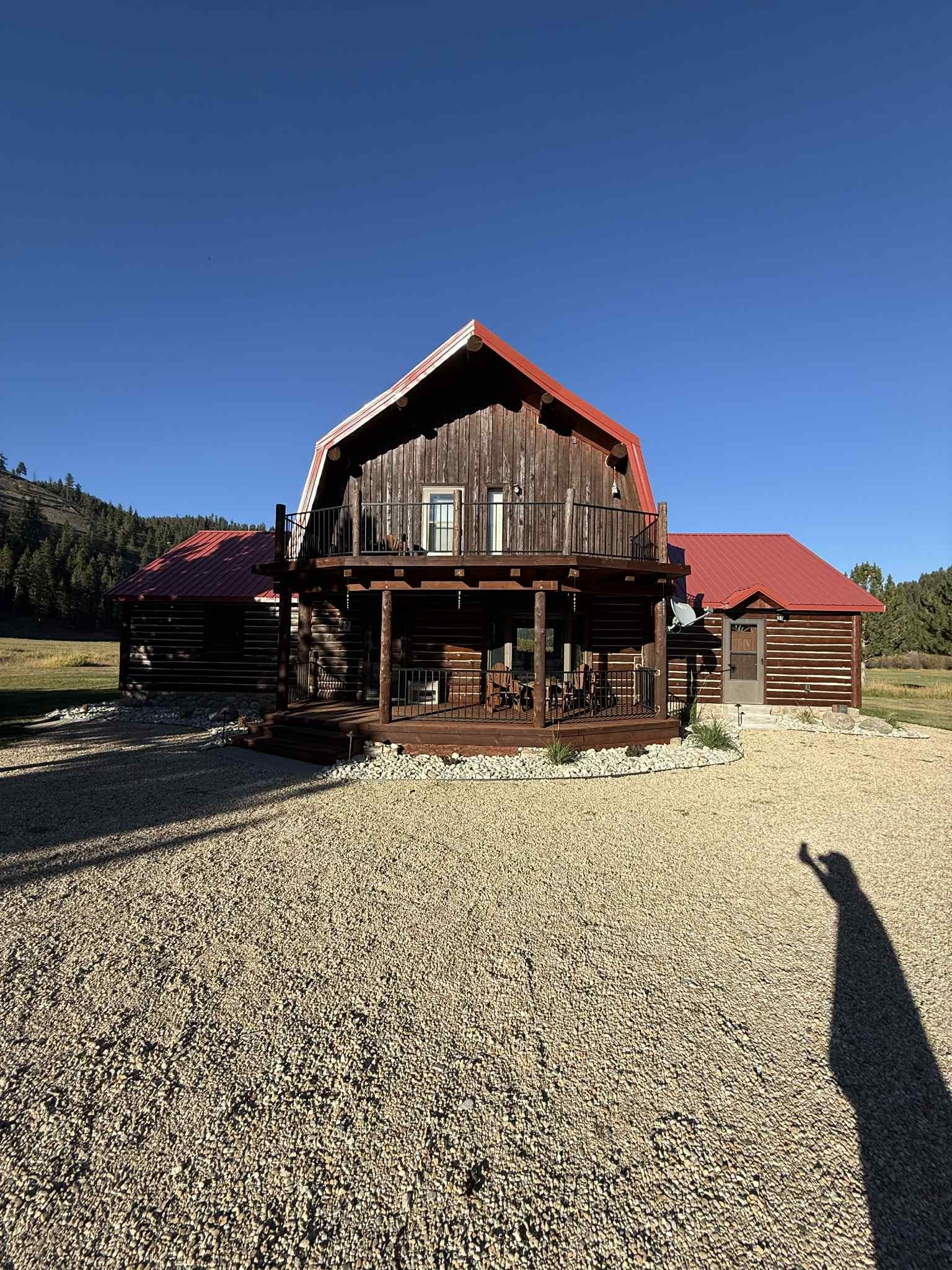 Rustic log cabin with a red roof, a porch, and a balcony on a gravel driveway under a blue sky.