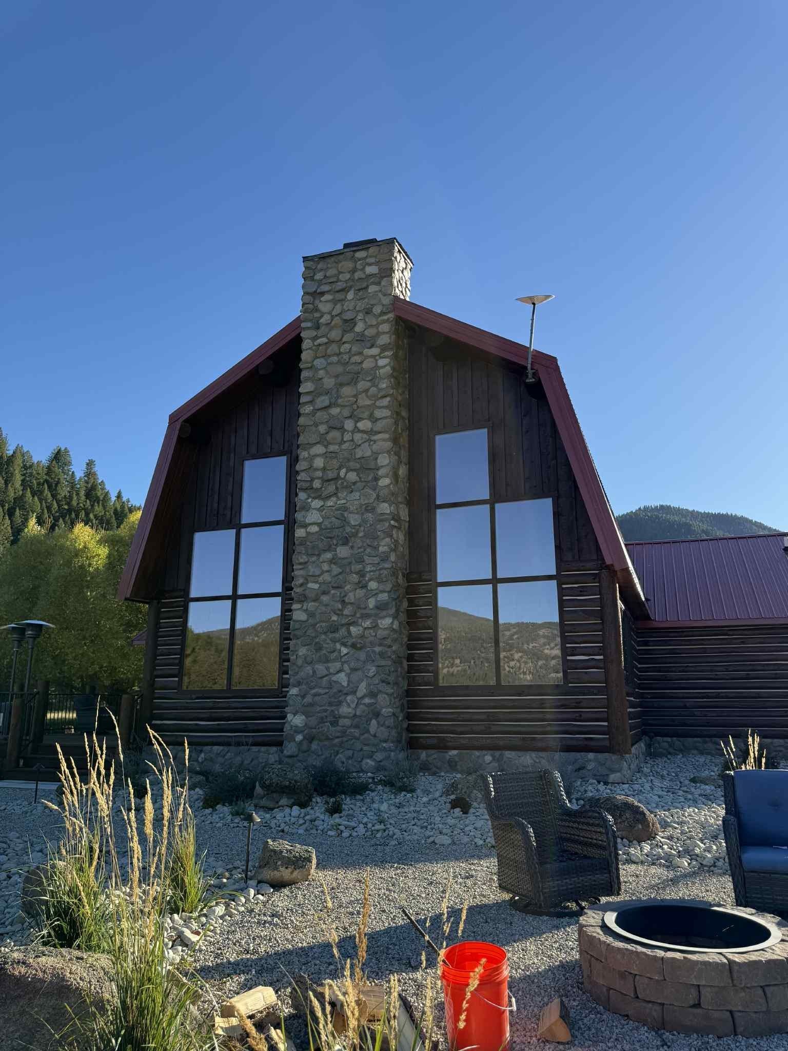 Log cabin with stone chimney, large windows, and a fire pit in a mountainous setting under a clear sky.