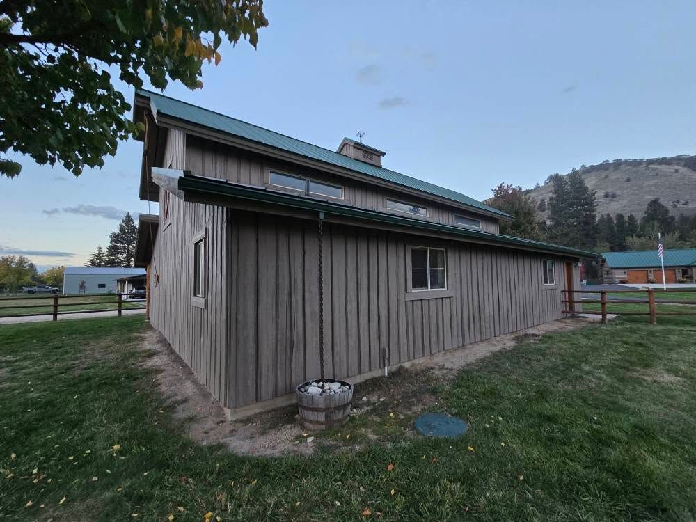 Gray wooden barn with green roof, windows, and rain gutters, on a grassy field; mountains in background.