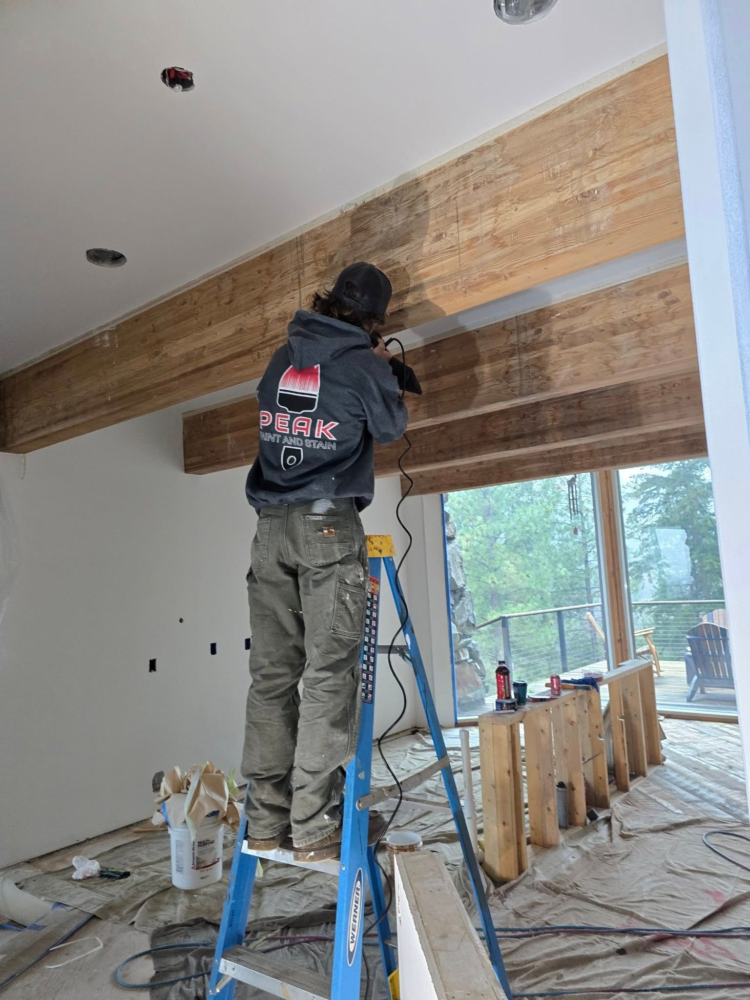 A worker on a ladder cuts into a wooden beam in a room under construction.