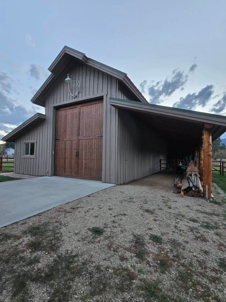 Barn with large wooden doors, tan siding, and a covered side entry. Gravel driveway.