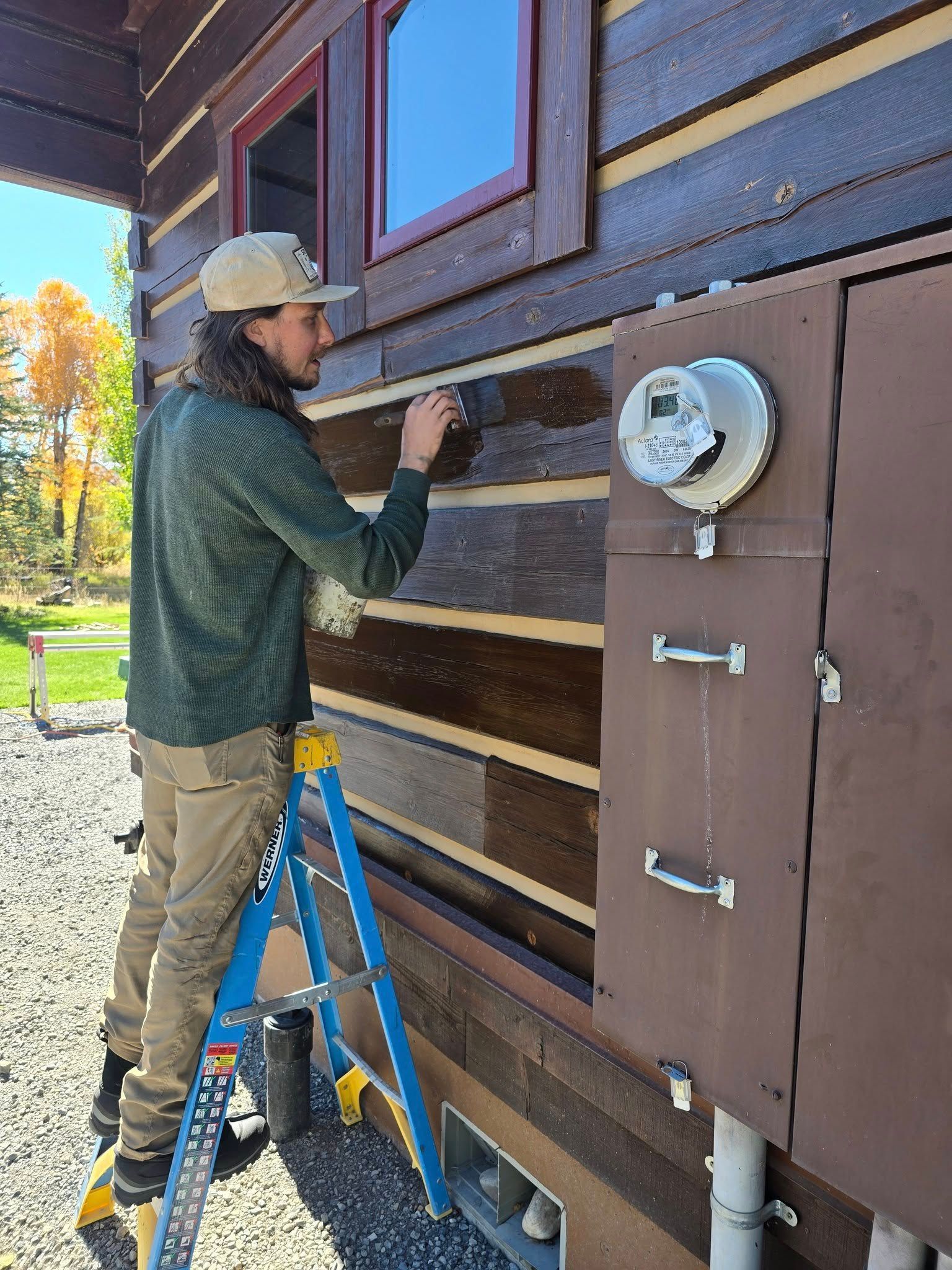 Person on ladder staining a log cabin exterior, next to electrical box.