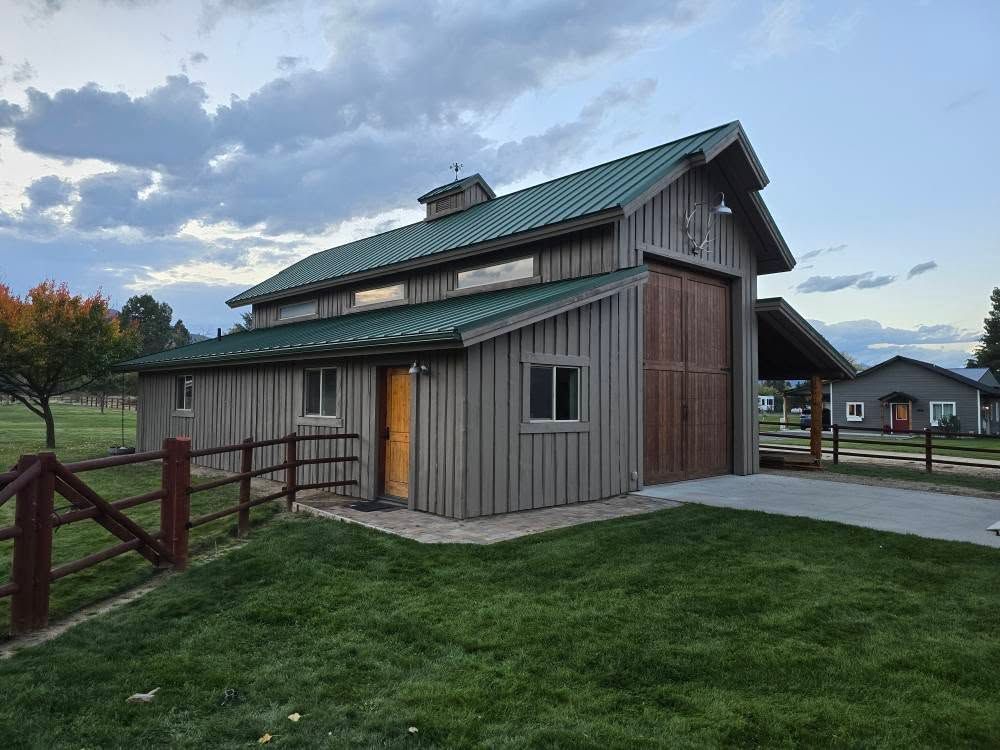 Wooden barn with green roof, open door, and adjacent grassy area under a cloudy sky.