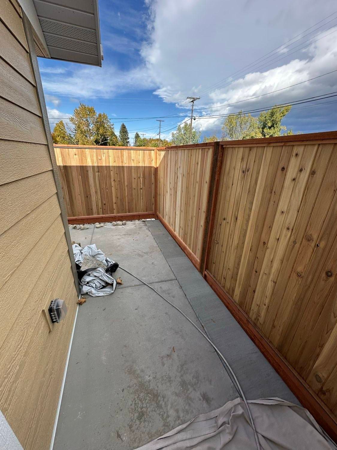 Concrete patio enclosed by a new wooden fence; beige siding on the left. Cloudy sky overhead.