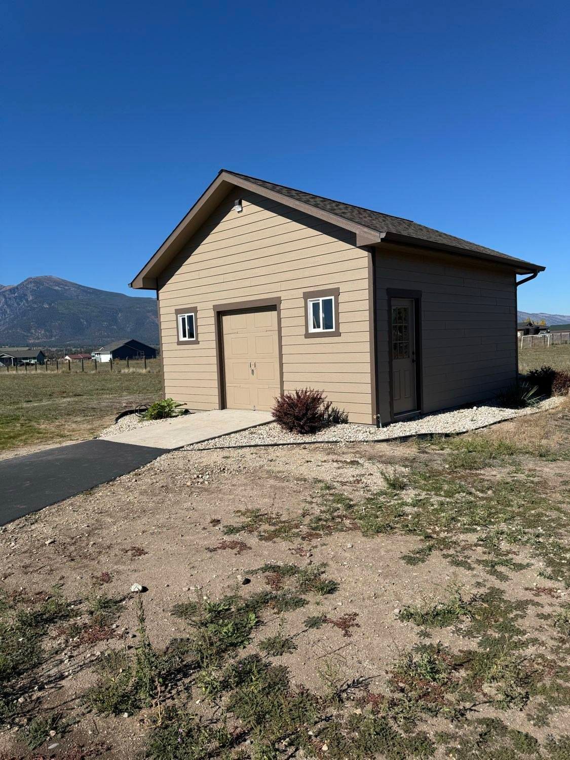 Tan building with garage door and small windows on a gravel lot under a blue sky, mountains in the distance.