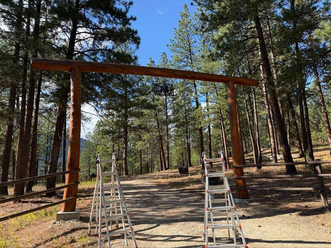 Wooden archway in a forest setting. Two ladders are in front. Sunny day with blue sky.